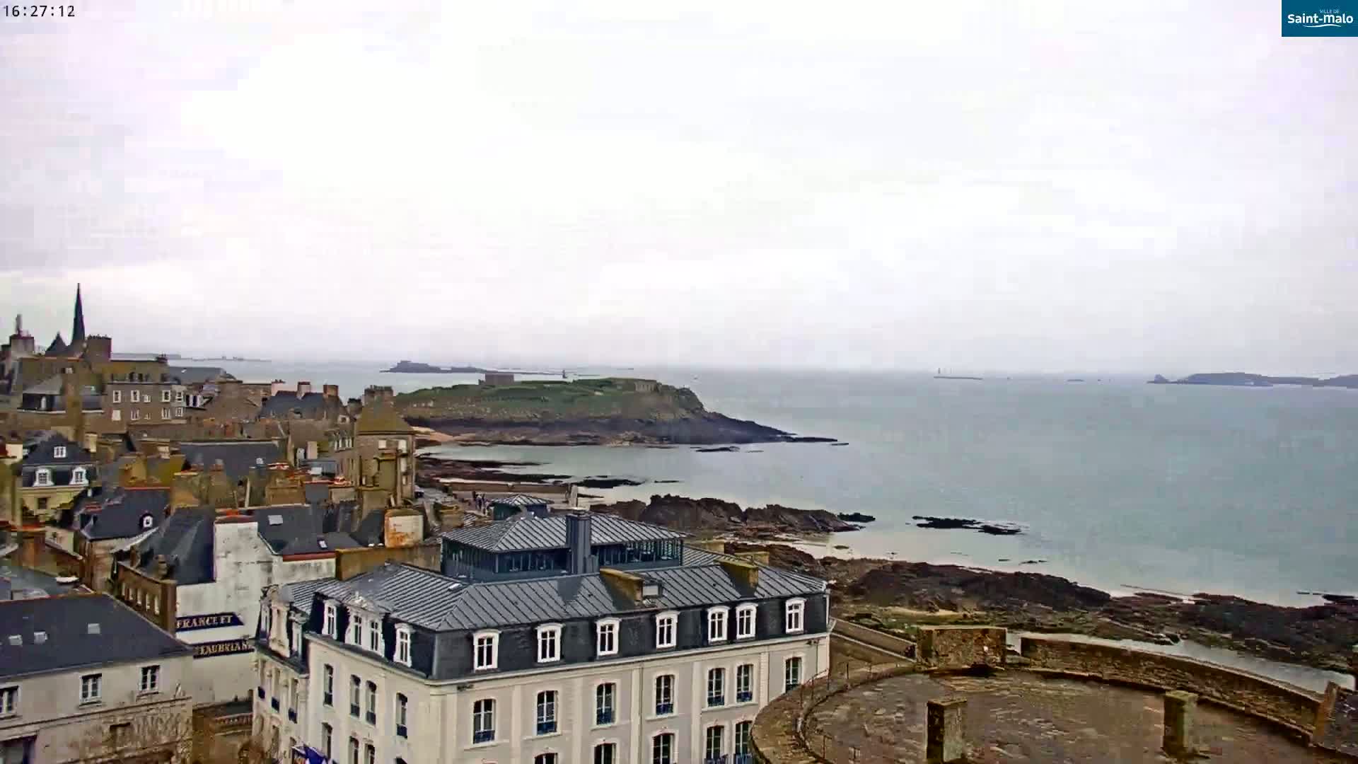 A coastal town with stone buildings overlooks a calm sea under an overcast sky.