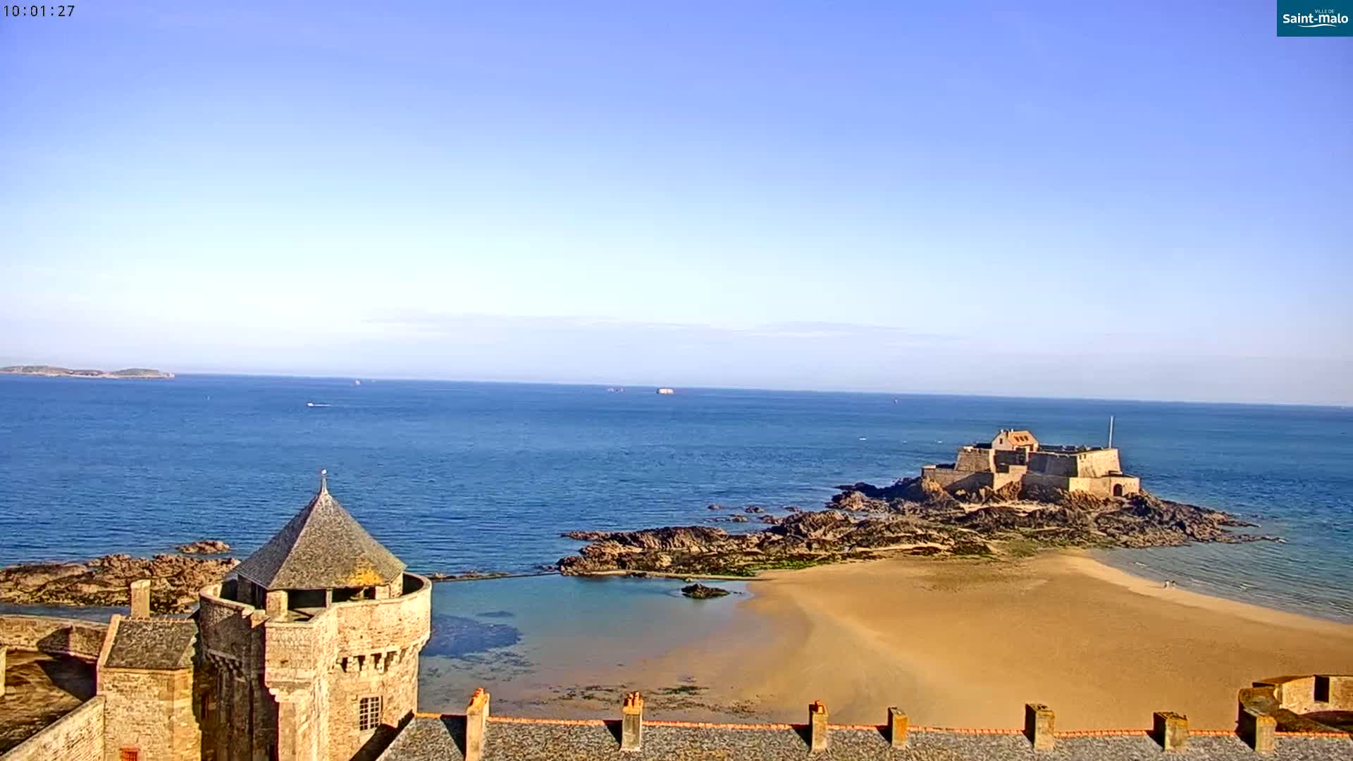 A coastal scene on a clear day shows a stone tower and fortress walls overlooking a sandy beach, a small body of water, rocky coastline, and a distant fortress on an island in the ocean.