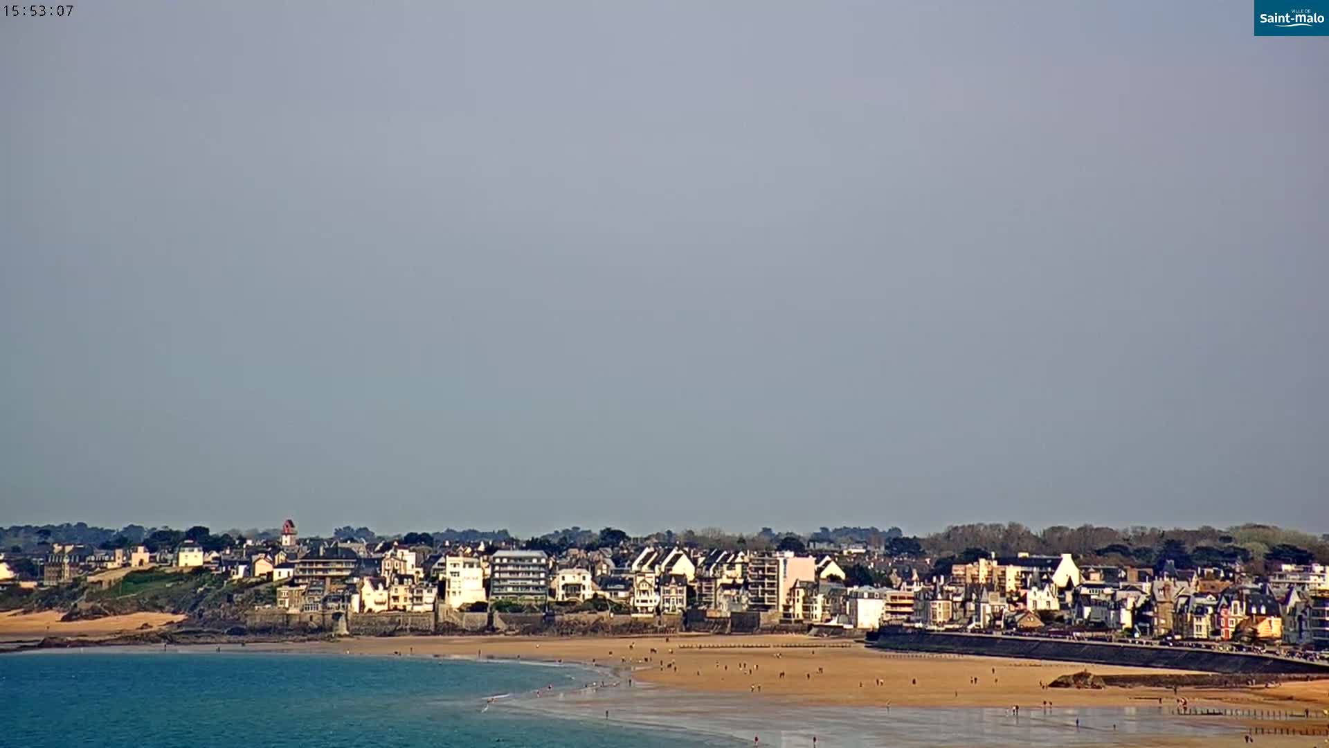 A coastal town with light-colored buildings sits on a sandy beach next to calm, blue water under a clear, light gray sky.