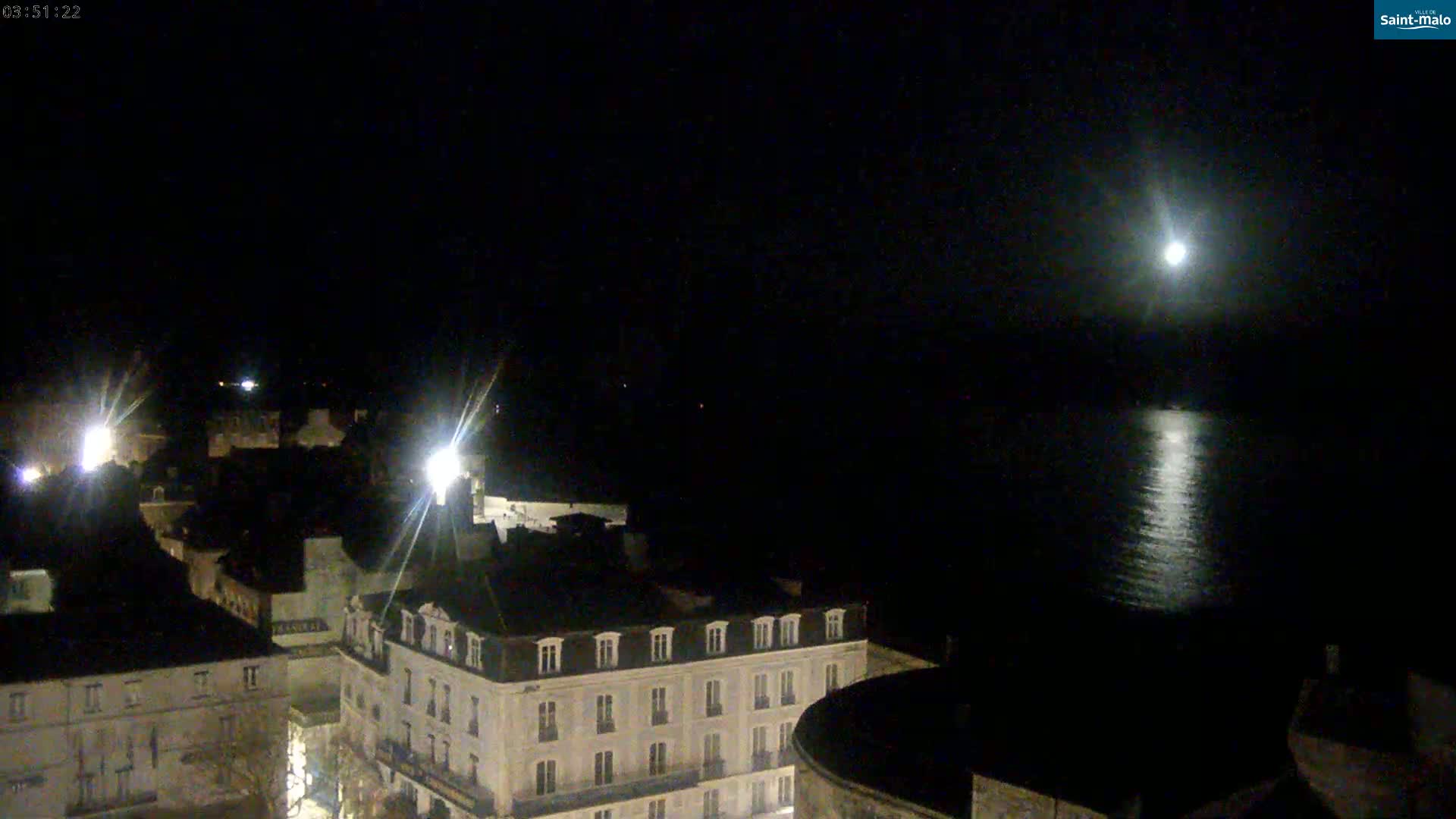 A nighttime view of a city waterfront illuminated by streetlights and a bright moon reflecting on the water.