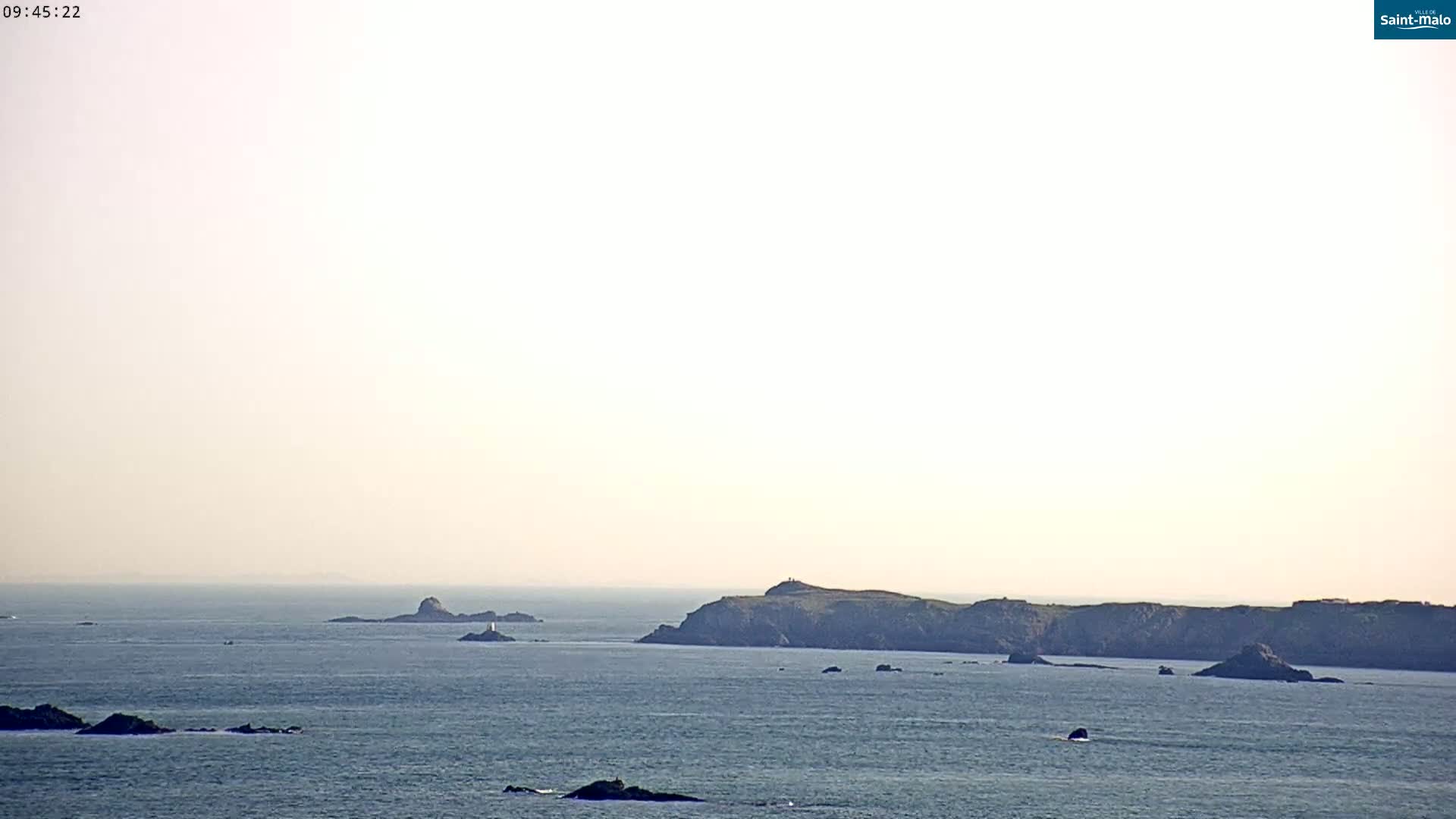 A hazy, calm ocean scene shows several dark, rocky islands and outcroppings under a pale sky.