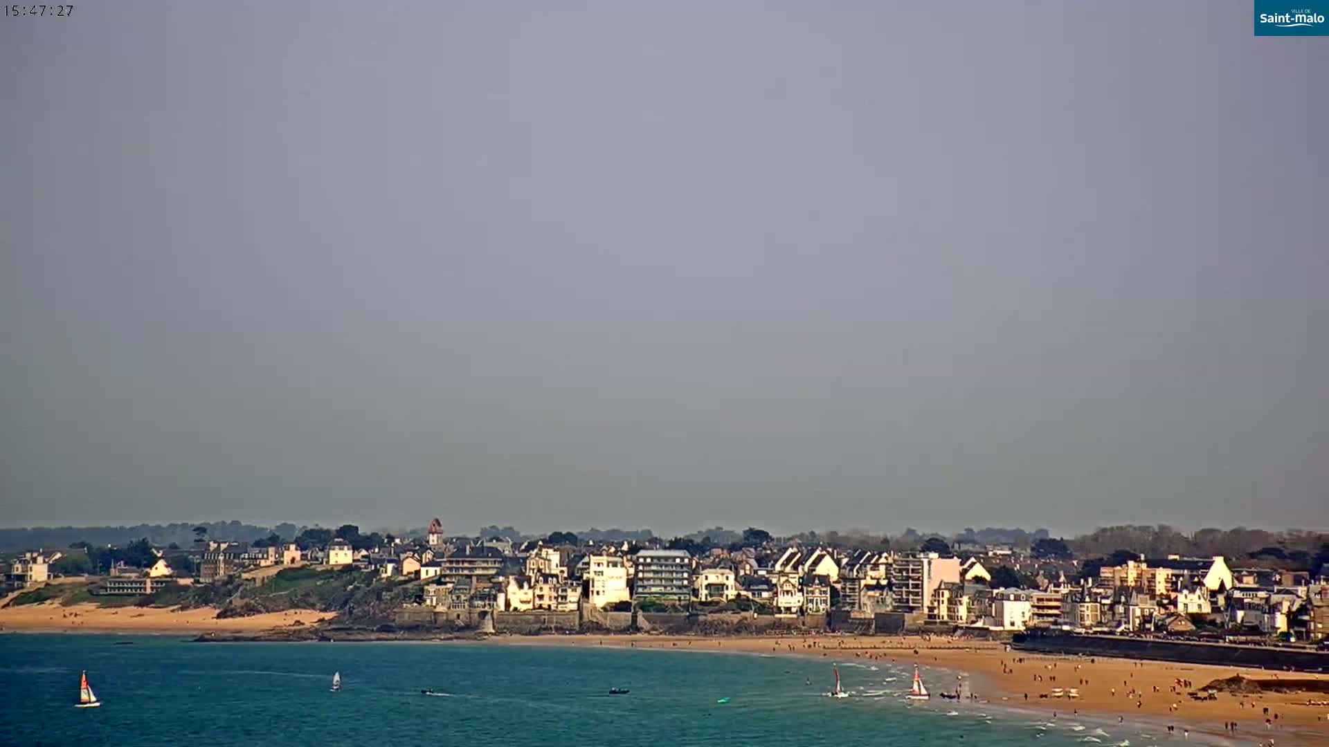 A hazy day reveals a coastal town with a sandy beach populated by people, and several sailboats on the calm ocean.