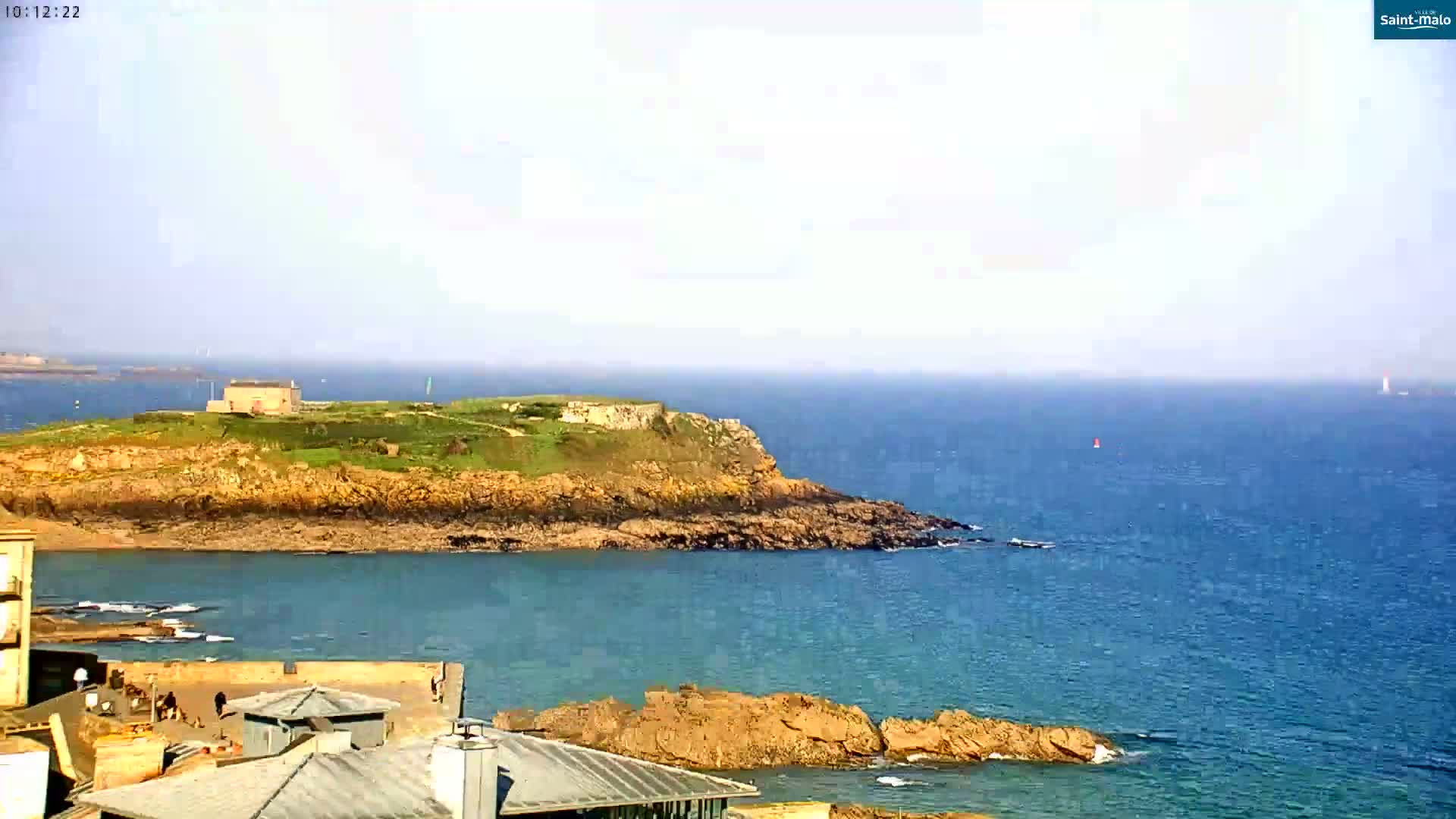 A hazy day shows a rocky coastal island with a building on top, surrounded by calm blue water and other rocky outcroppings.