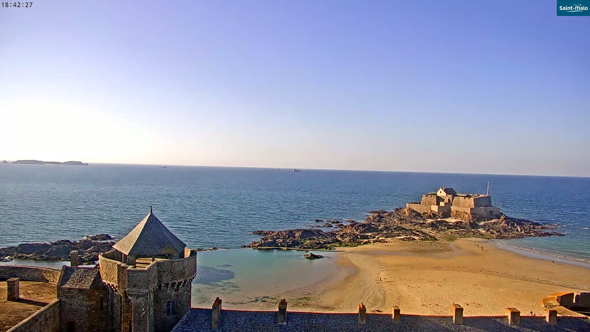 A stone castle overlooks a sandy beach, a small tidal pool, and a distant fortress on a rocky islet, all under a clear, sunny sky.