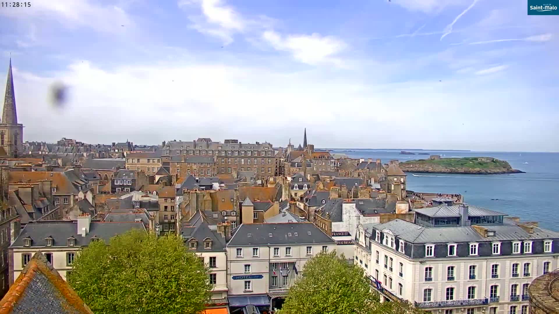 A coastal city with numerous stone buildings, a church steeple, and a small island in the distance under a partly cloudy sky.