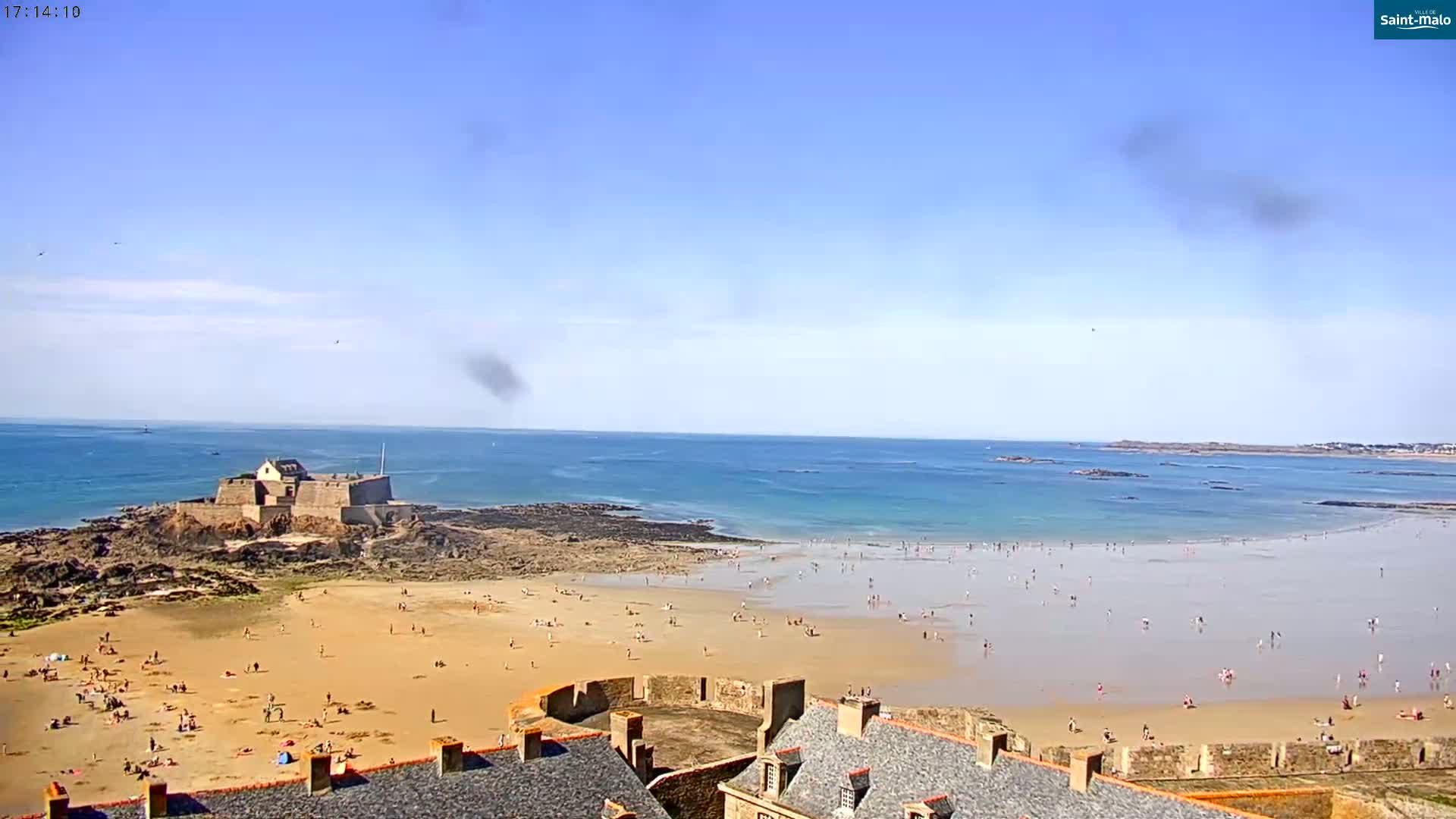 A sandy beach with many people on it stretches out from a stone fortress on a rocky outcrop to a calm ocean under a mostly clear blue sky.