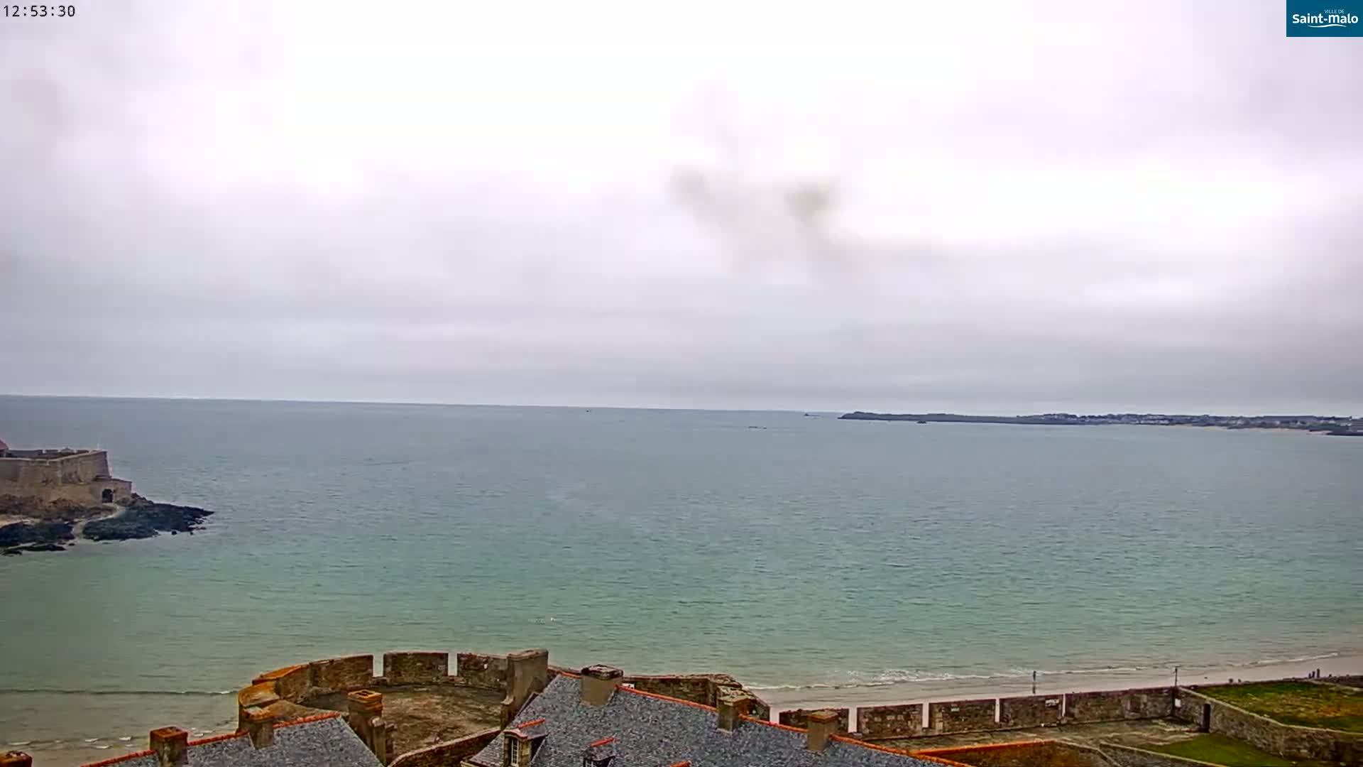 A coastal scene under an overcast sky shows a stone fortress overlooking a calm sea and a sandy beach.