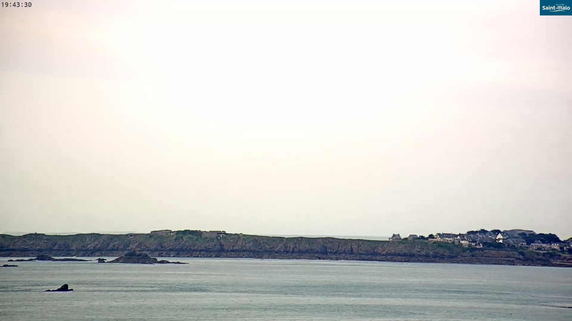 A rocky coastline with a small village on a distant island is visible under a hazy, overcast sky over calm water.