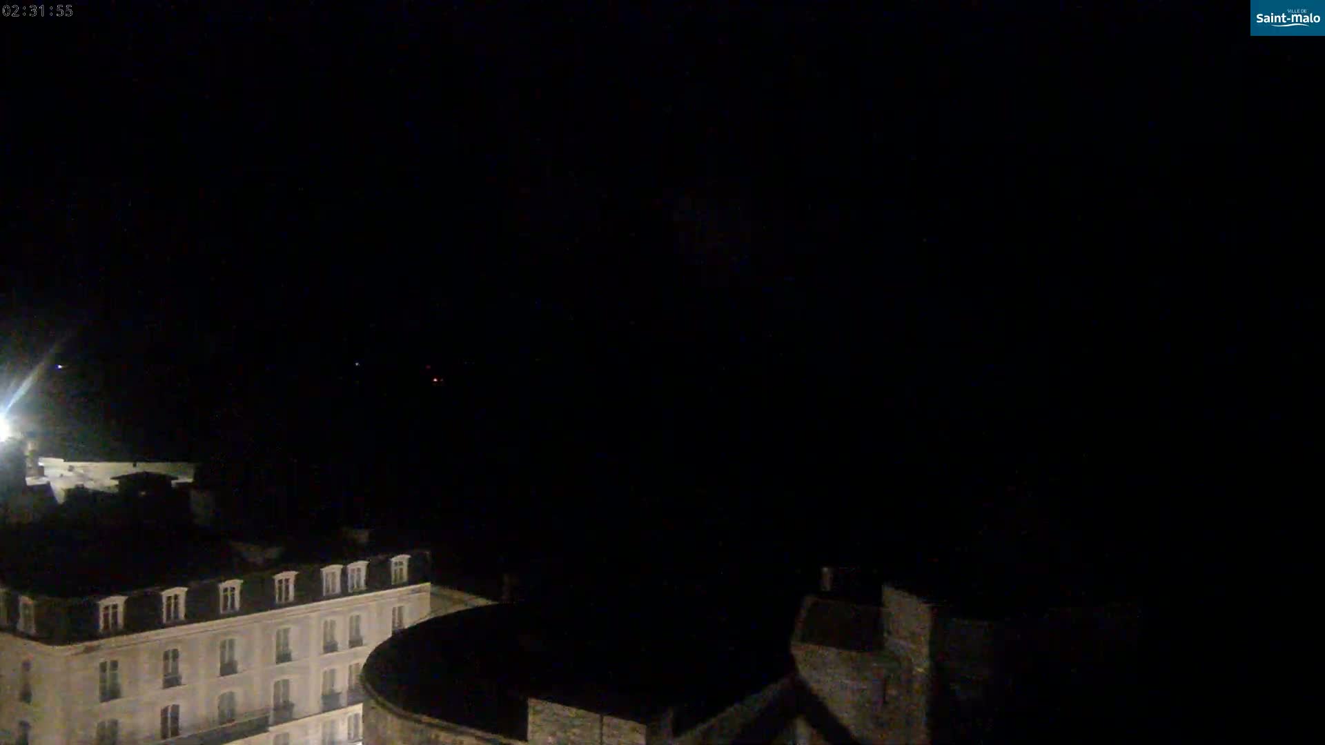 A nighttime, low-light view shows a section of a city with a few dimly lit buildings, under a dark, clear sky.