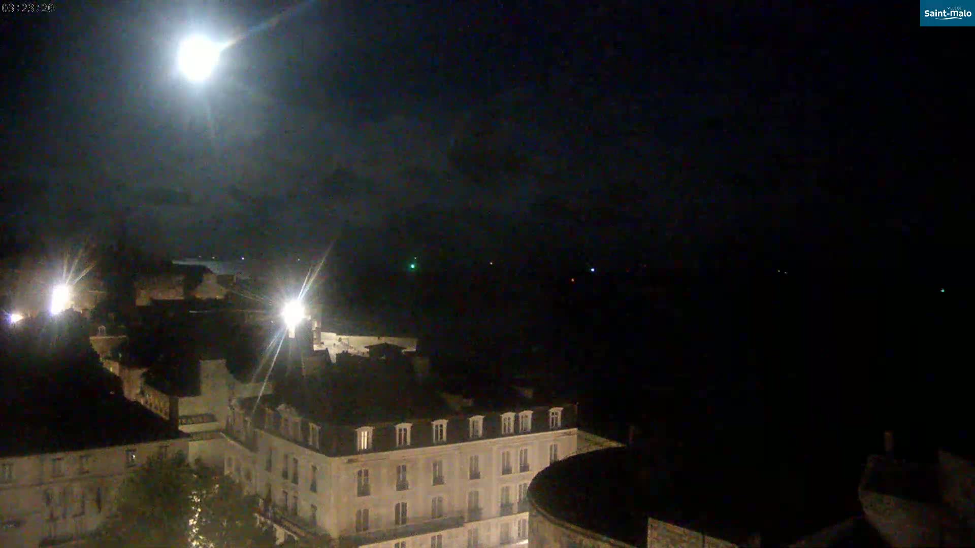 A bright moon illuminates a nighttime cityscape of buildings, with a dark body of water visible in the distance under a mostly clear sky.