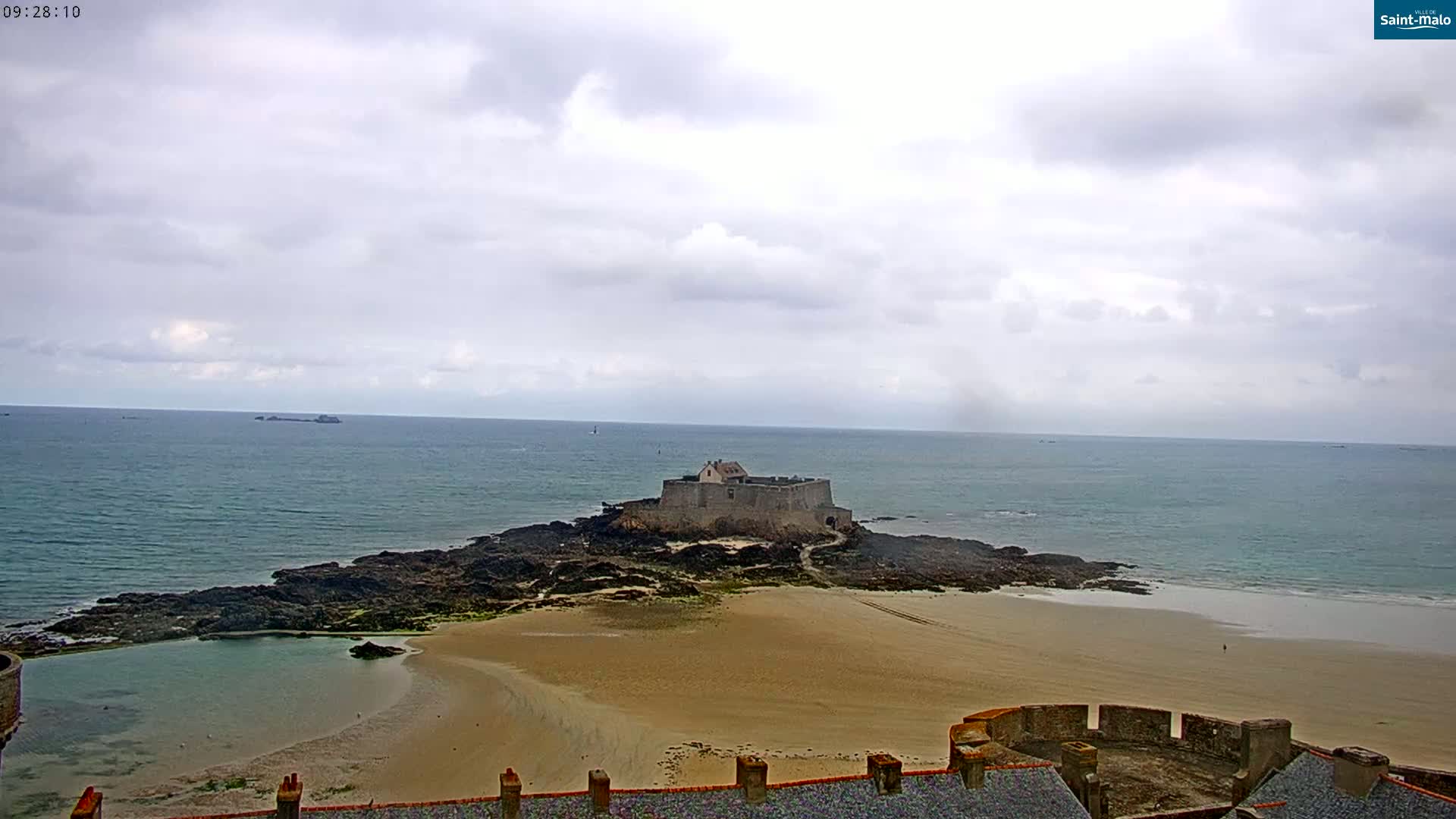 A stone fortress sits on a small island connected to a sandy beach by a rocky causeway, under a cloudy sky.