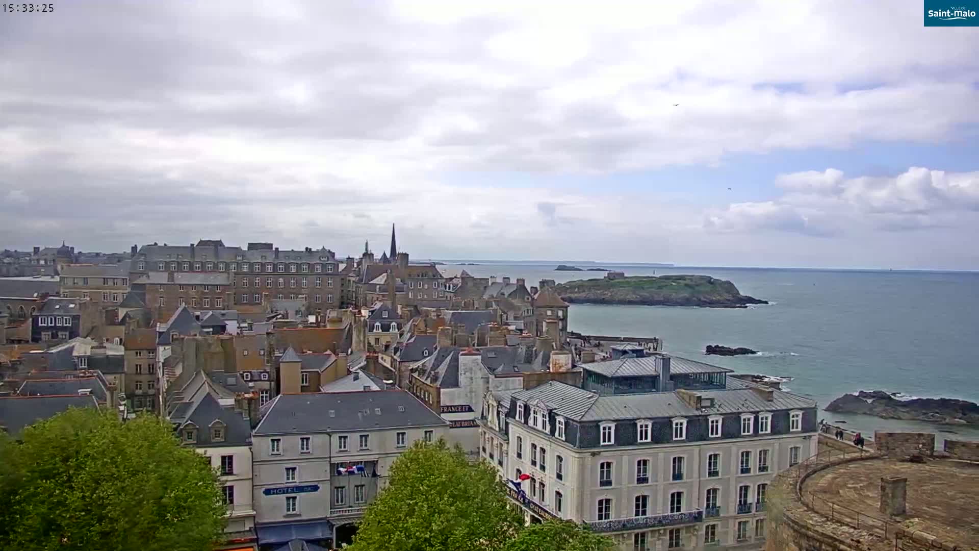 A coastal city with many stone buildings is shown under a partly cloudy sky, with a calm ocean in the background.
