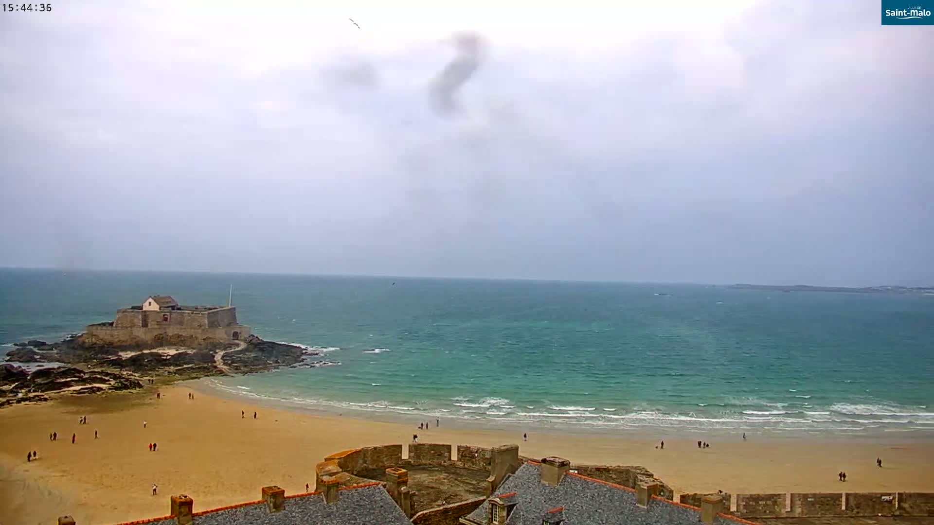 A sandy beach with a small fort on an island in the ocean, under an overcast sky.