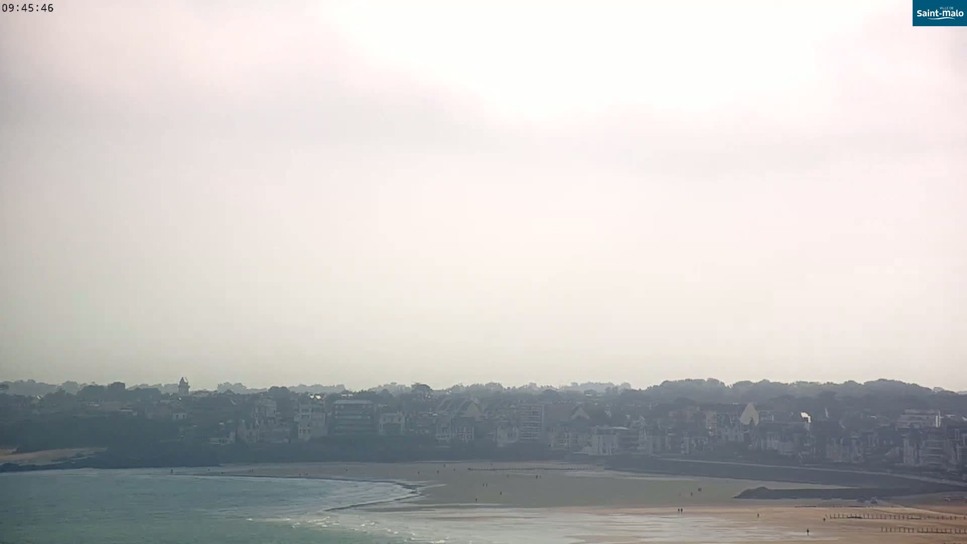 A hazy coastal town with a sandy beach and calm ocean is visible from a distance.