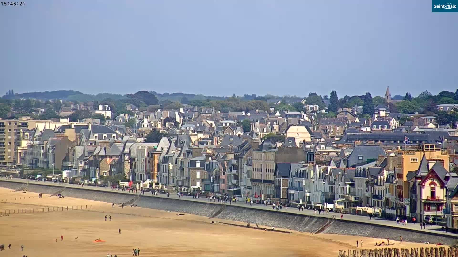 A sandy beach with people walking along a seawall and numerous closely-packed buildings extending into the background under a clear sky.