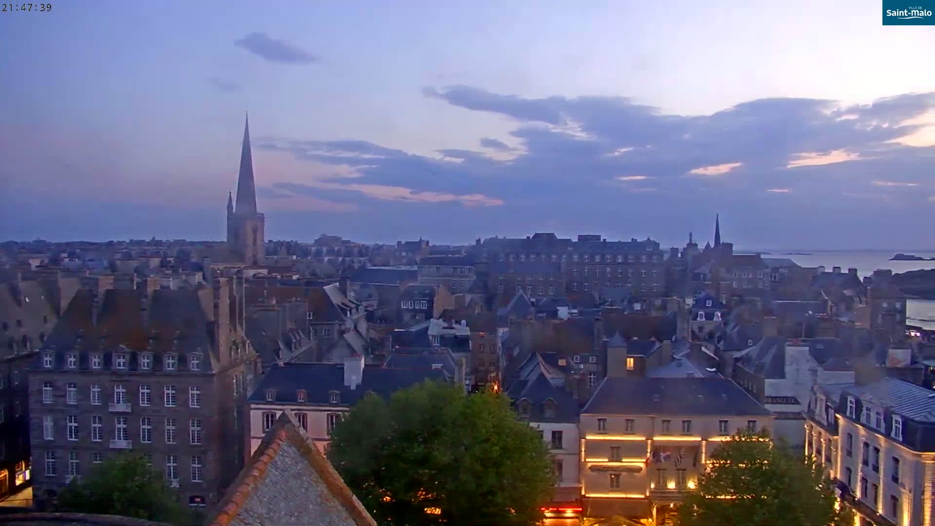 A twilight view of a coastal town shows numerous buildings, a tall church spire, and calm water in the distance under a partly cloudy sky.