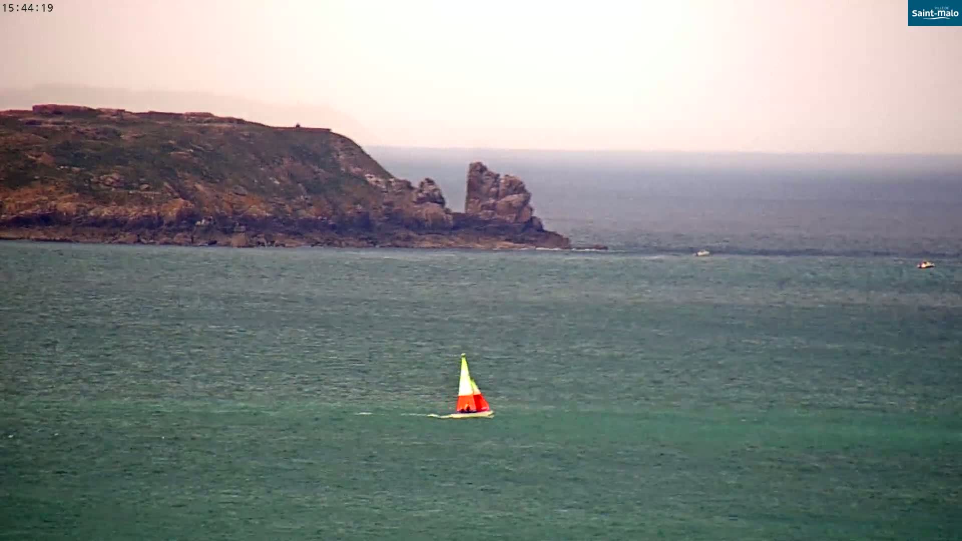 A small sailboat with an orange and yellow sail is on a body of dark green water in front of a rocky coastline under an overcast sky.