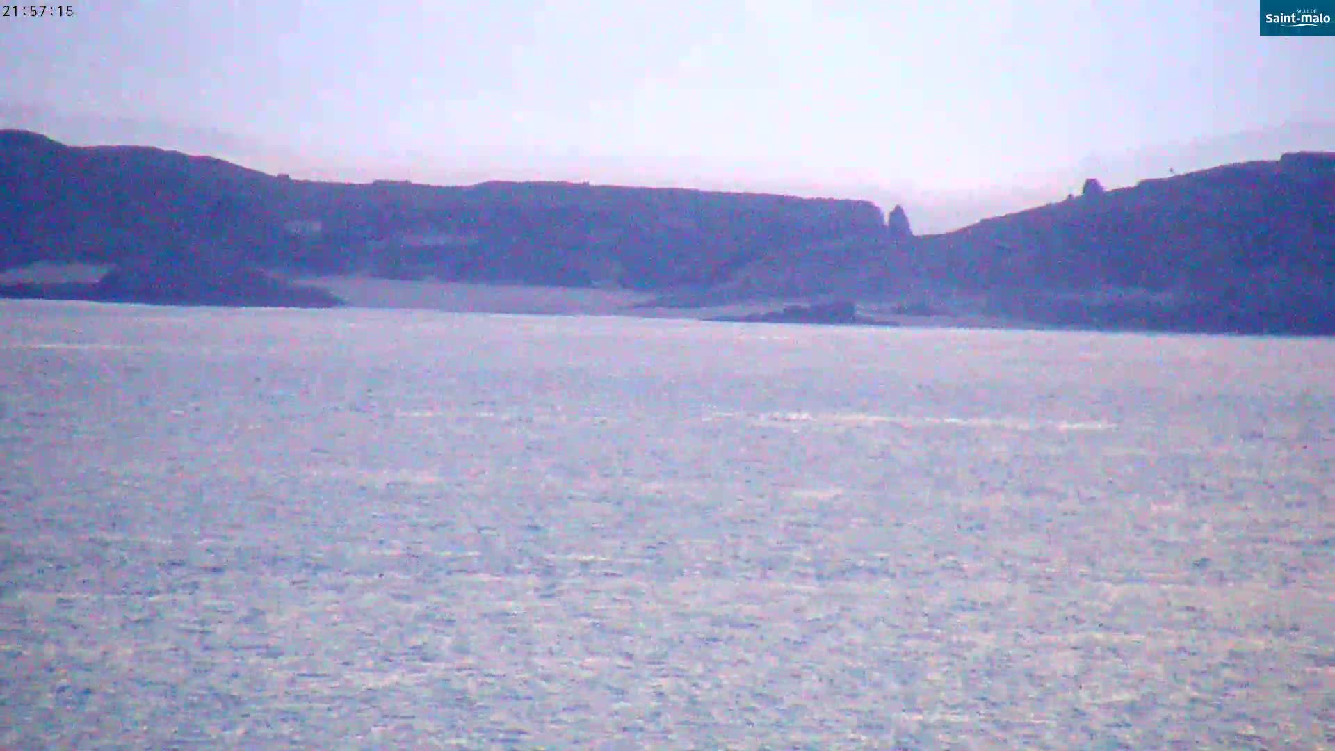 A calm body of water stretches out before a distant, dark, hilly coastline under a pale sky.