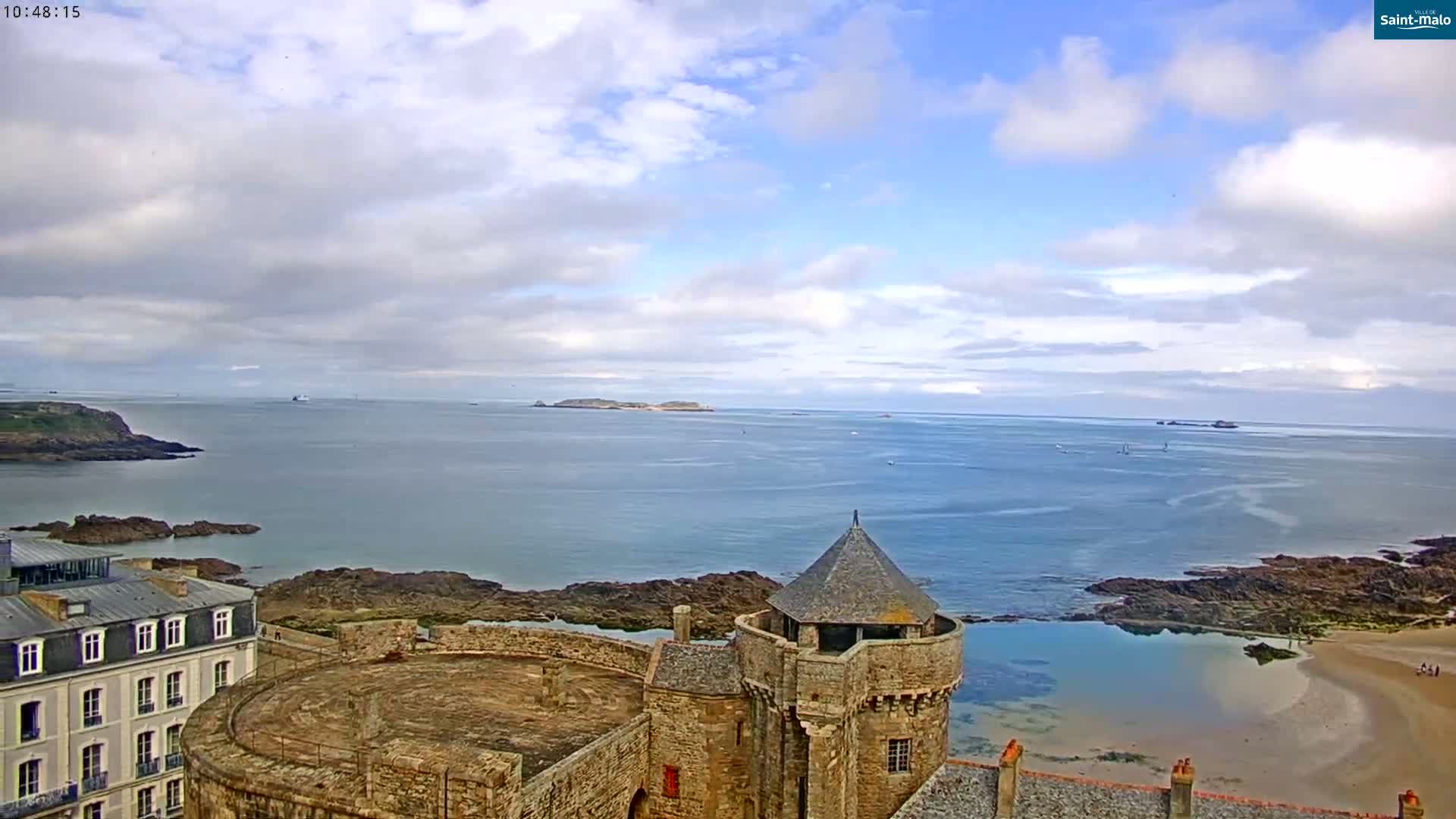 A stone tower overlooks a calm ocean bay with rocky shores, a sandy beach, and distant islands under a partly cloudy sky.
