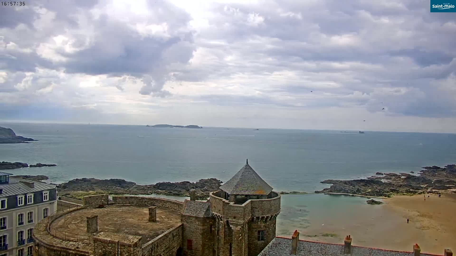 A stone coastal fortress overlooks a calm ocean under a cloudy sky, with a small sandy beach visible below.