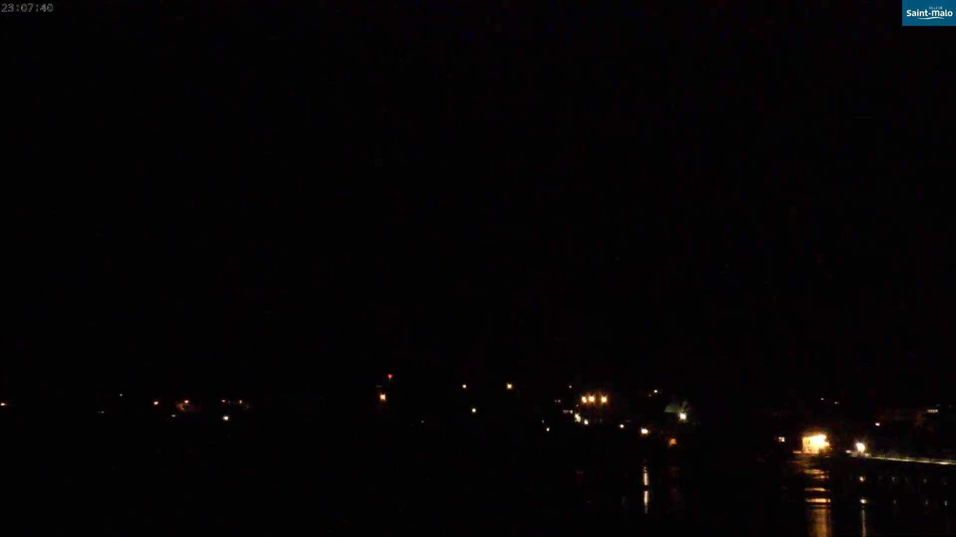 A nighttime view of a dark body of water reflecting scattered, distant lights from a shoreline under a clear, dark sky.