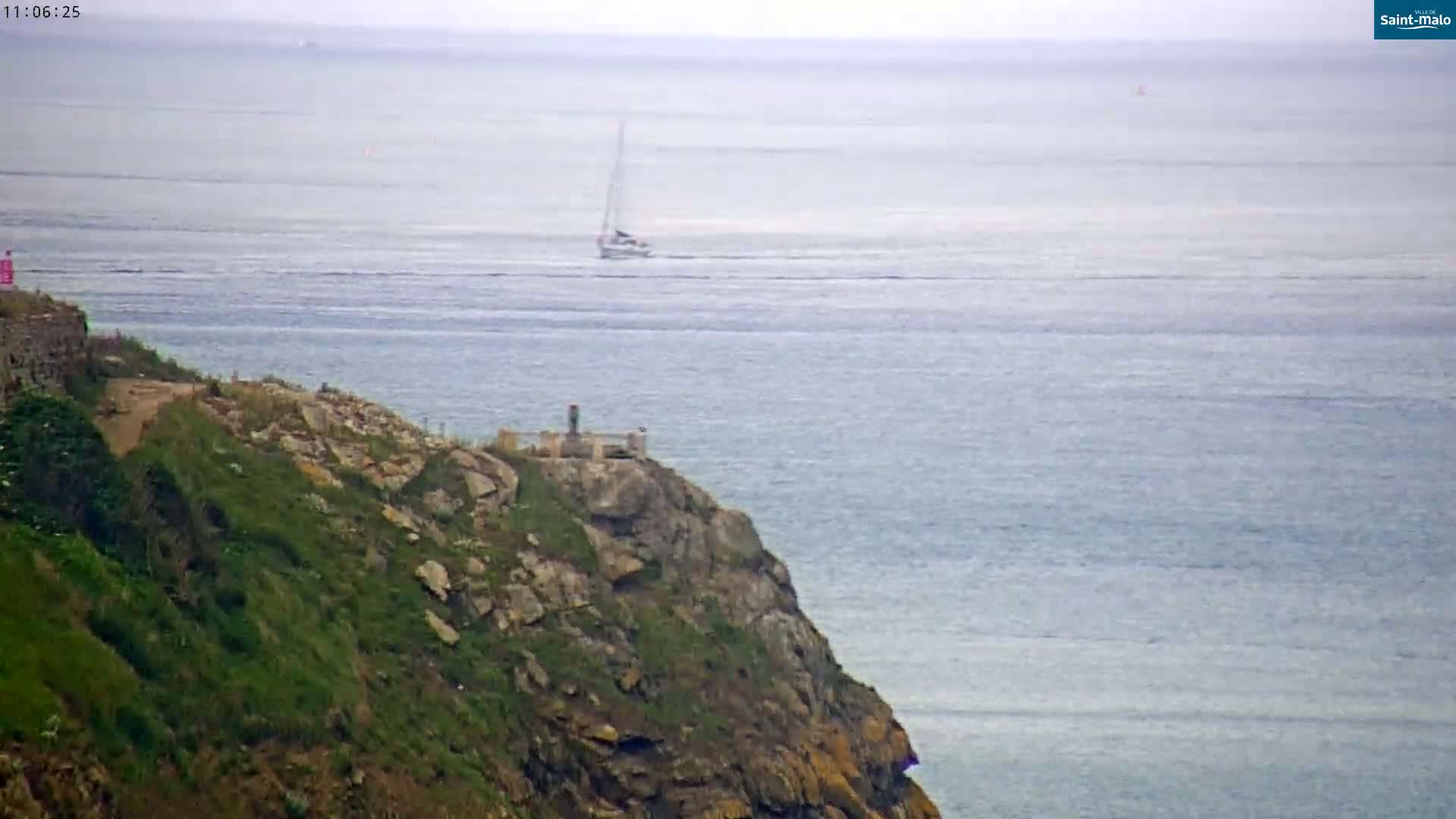 A sailboat is visible on a calm, gray ocean from a rocky, grassy cliff.
