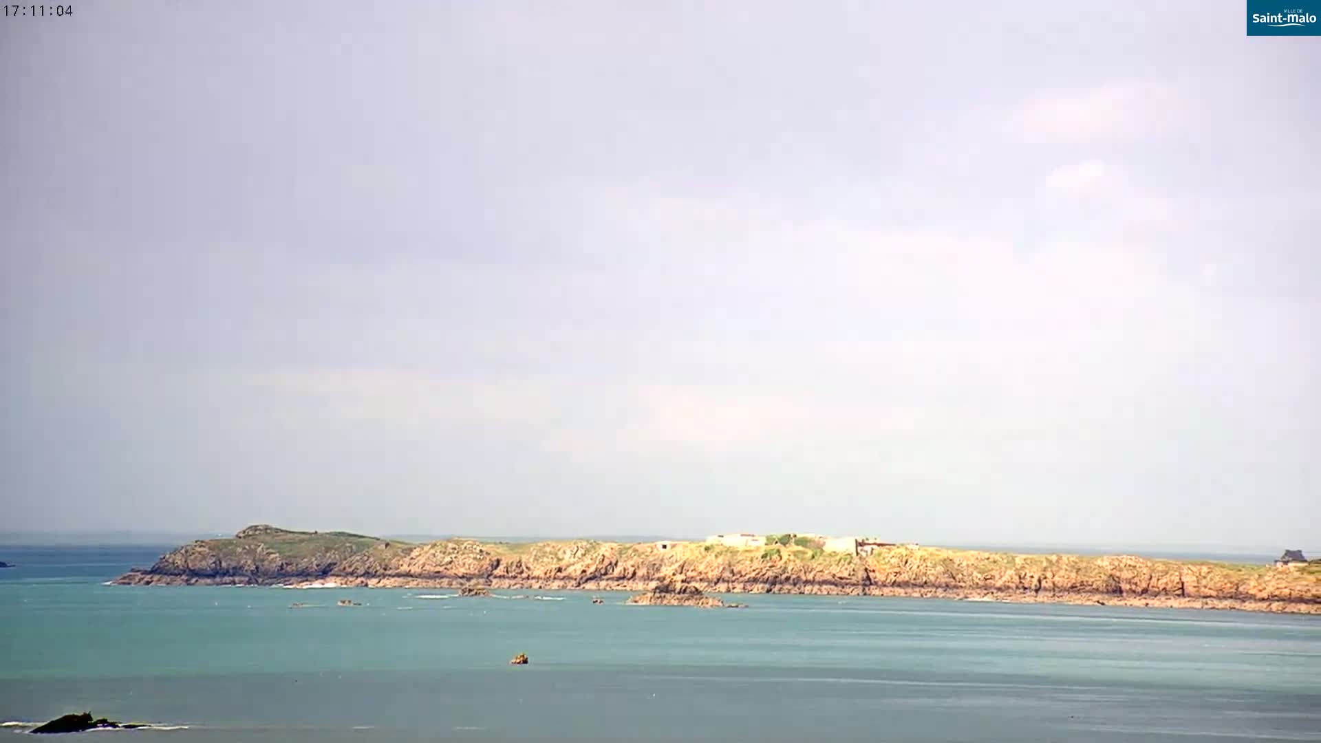 A rocky island stretches across a calm, light teal sea under a mostly cloudy, light-grey sky.