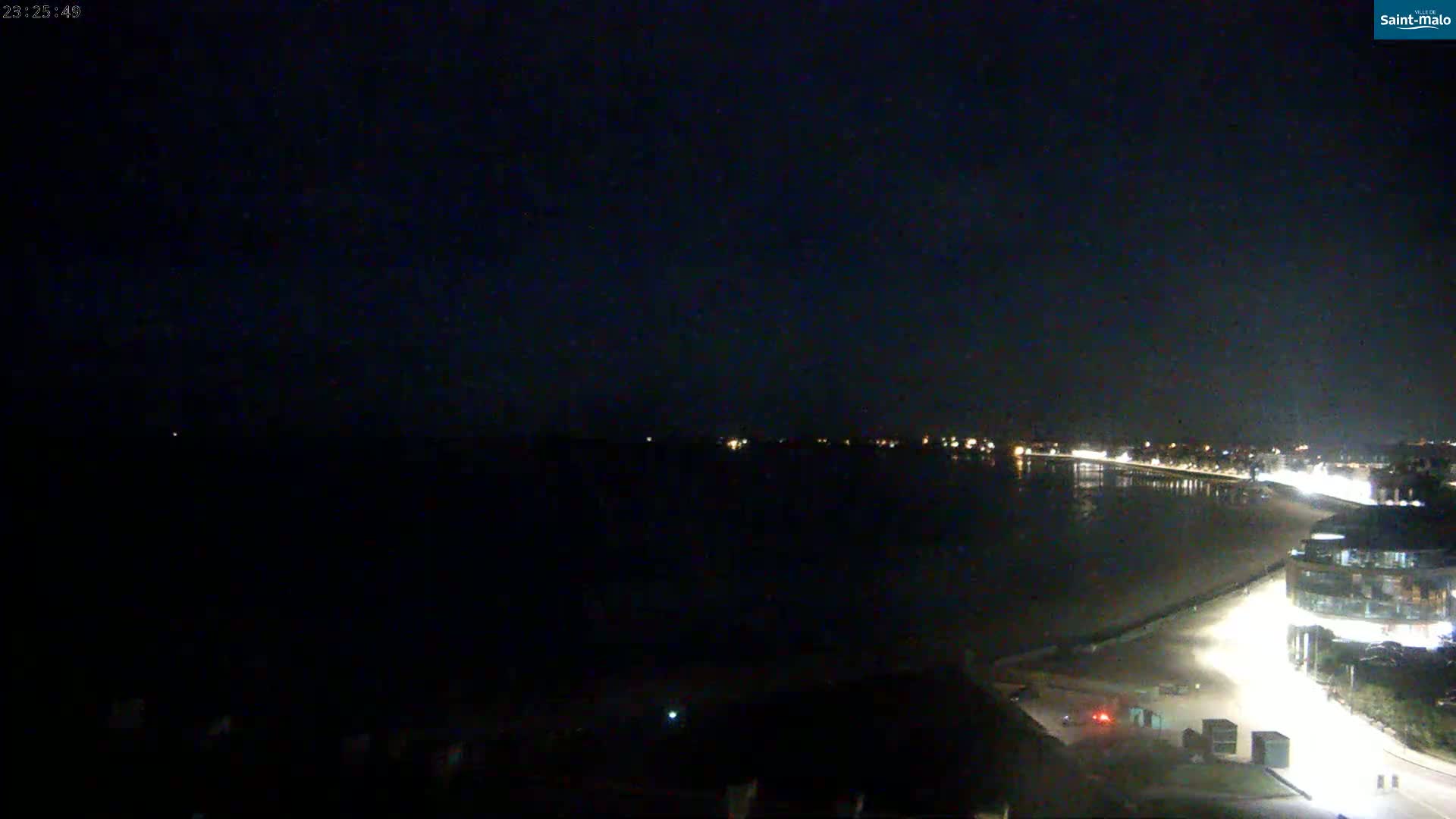 A nighttime view of a dark, calm body of water with a brightly lit beachfront and buildings visible in the distance under a clear, dark sky.