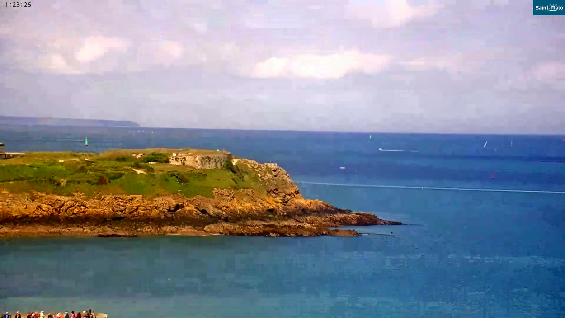 A rocky, green-topped headland juts into a calm, blue ocean under a partly cloudy sky.