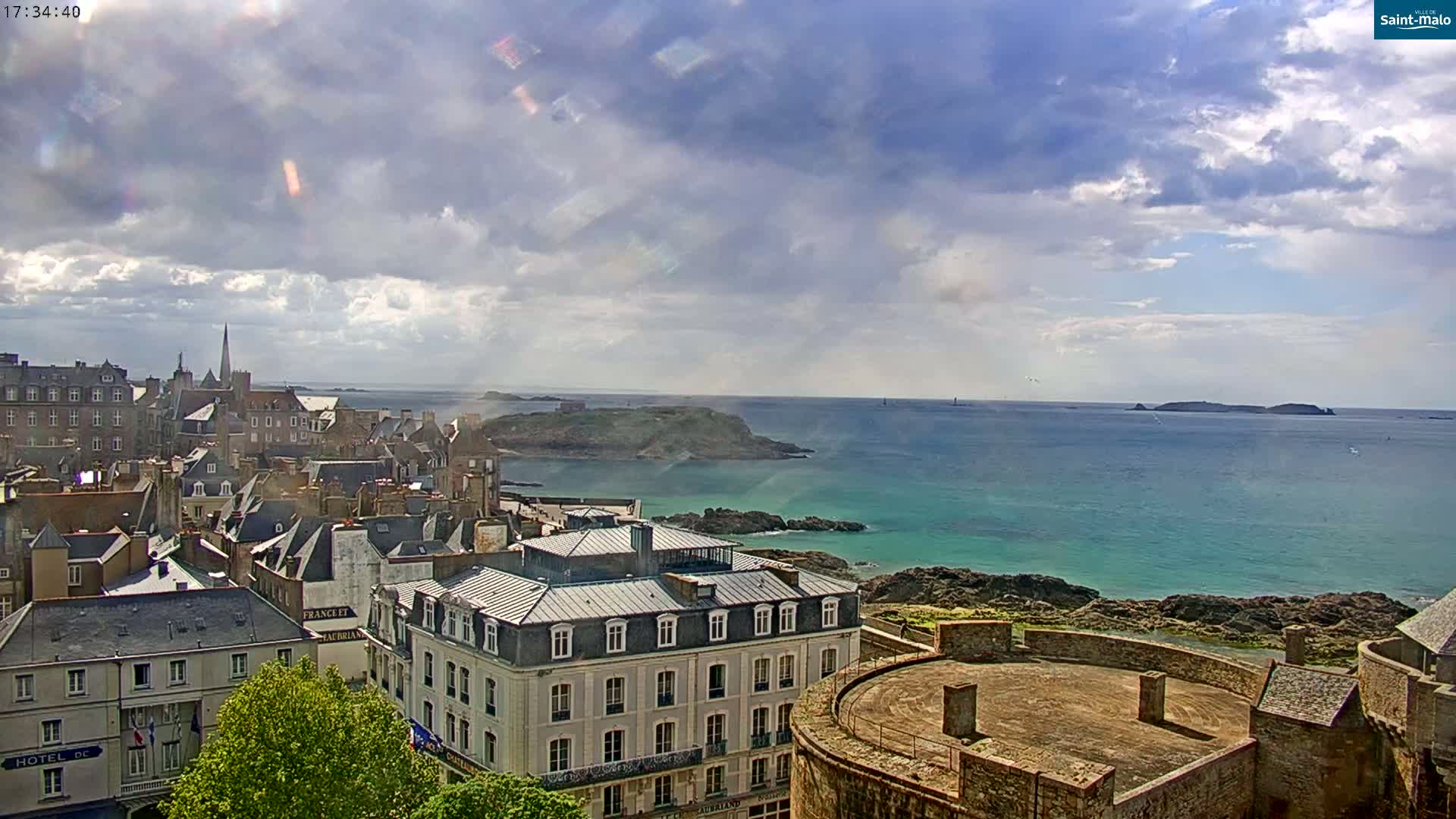 A coastal city with stone buildings and a partially cloudy sky overlooks a turquoise ocean and small islands.