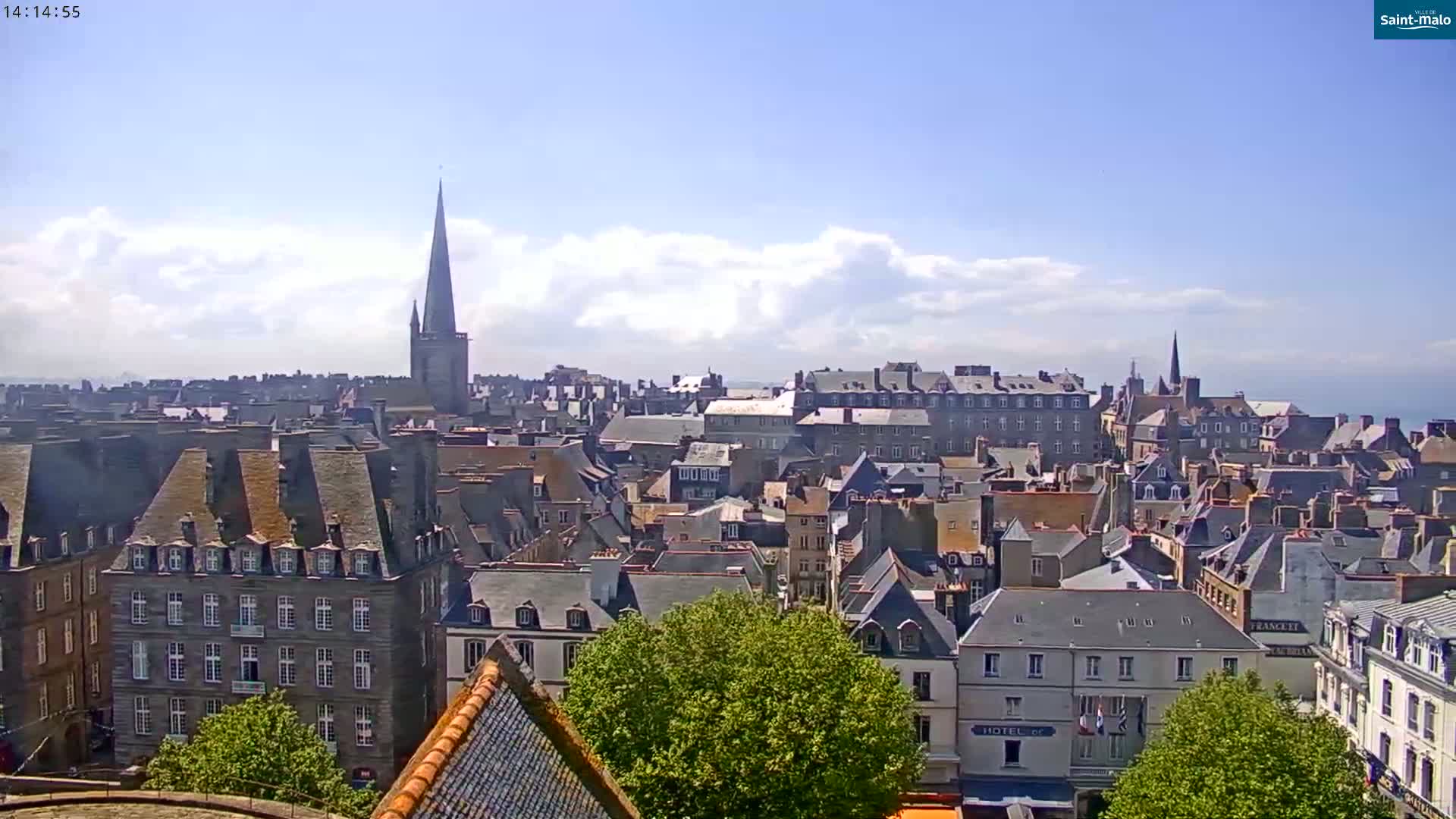 A high-angle view of a European city on a partly cloudy, sunny day, showing many grey-roofed buildings and a tall church spire.