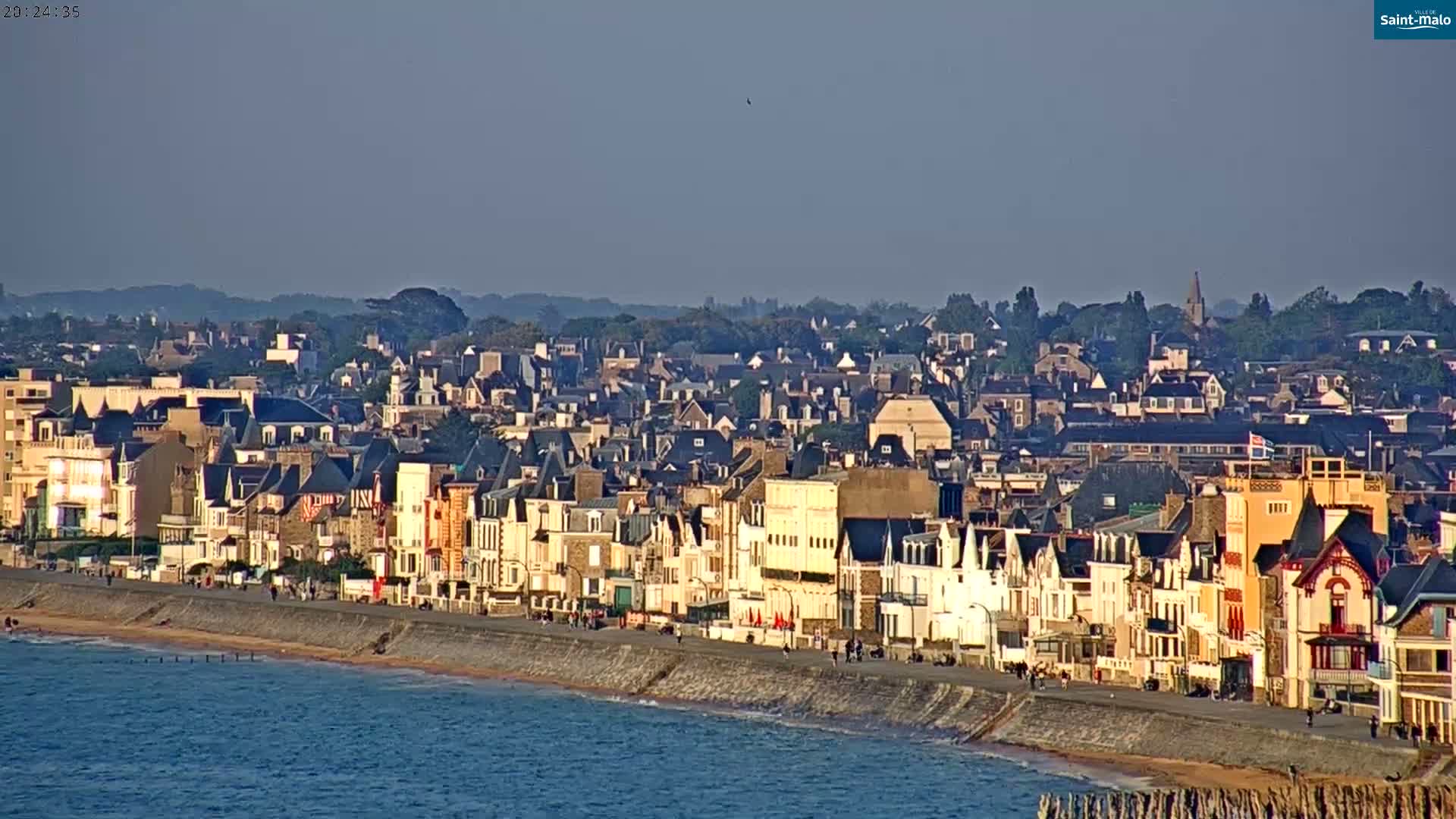 A coastal town with many buildings sits on a sandy beach next to calm, blue water under a clear, light-blue sky.