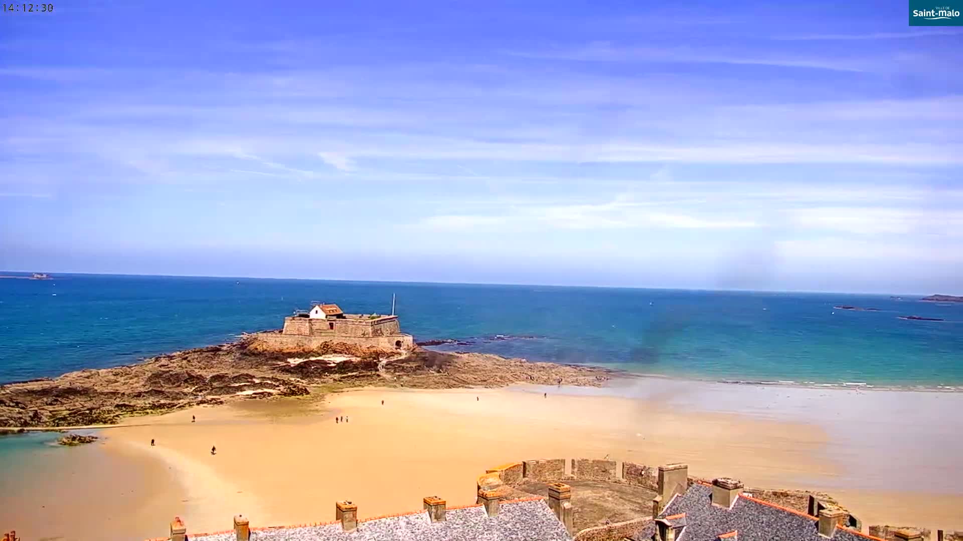 A stone fort sits on a rocky outcrop beside a sandy beach and calm, blue ocean under a mostly sunny sky.
