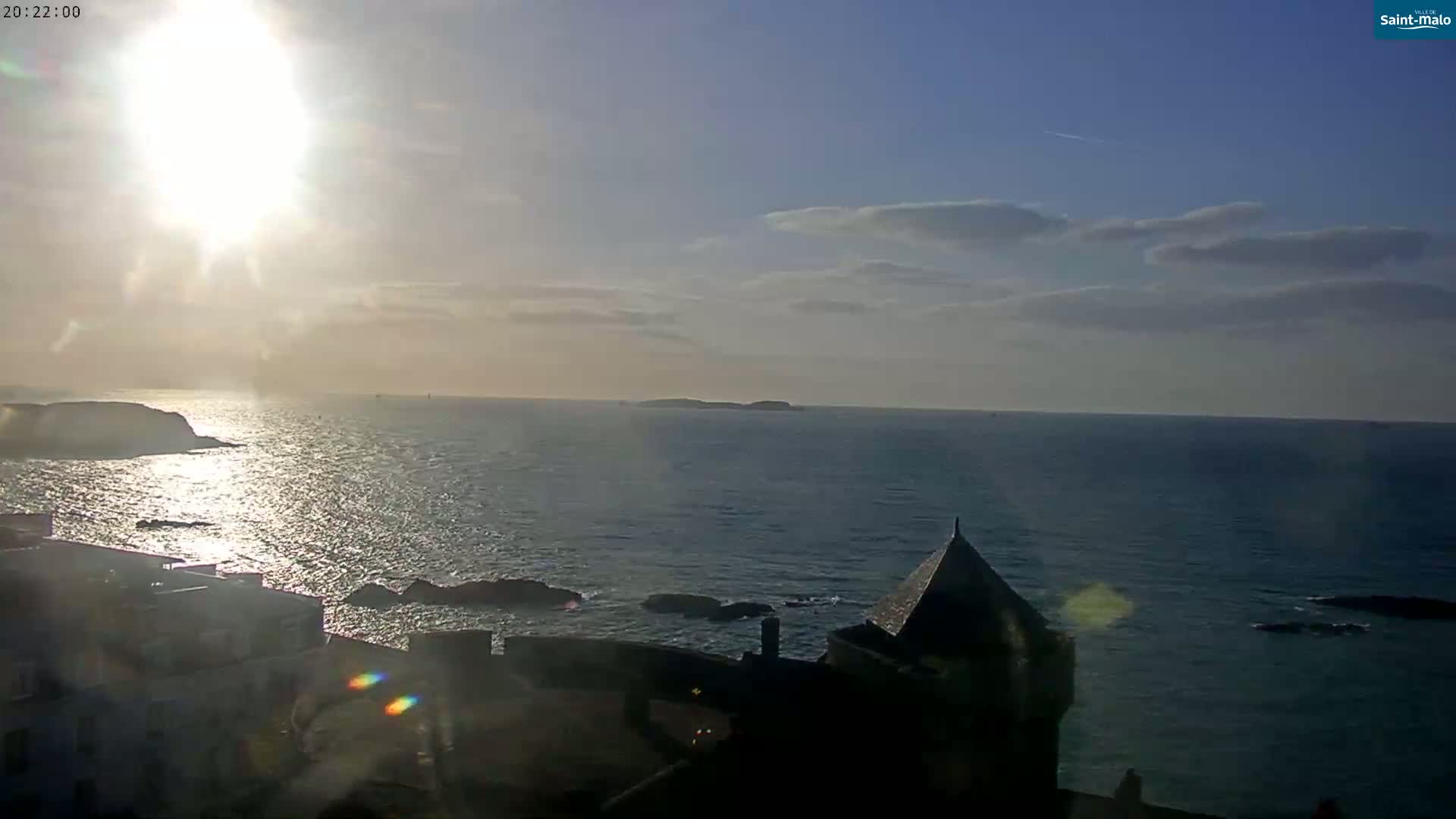 A bright sun shines over a calm ocean, illuminating a coastal town with a stone structure near the water.