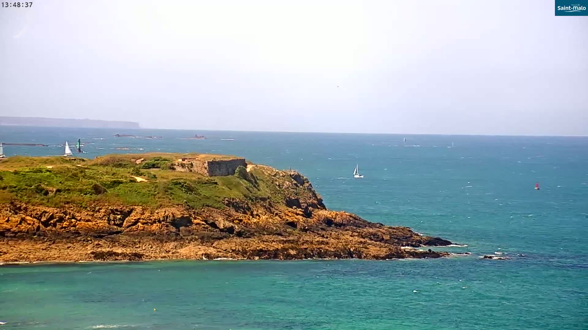 A rocky, grassy headland juts into a calm, turquoise sea under a clear, bright sky, with several sailboats visible in the distance.
