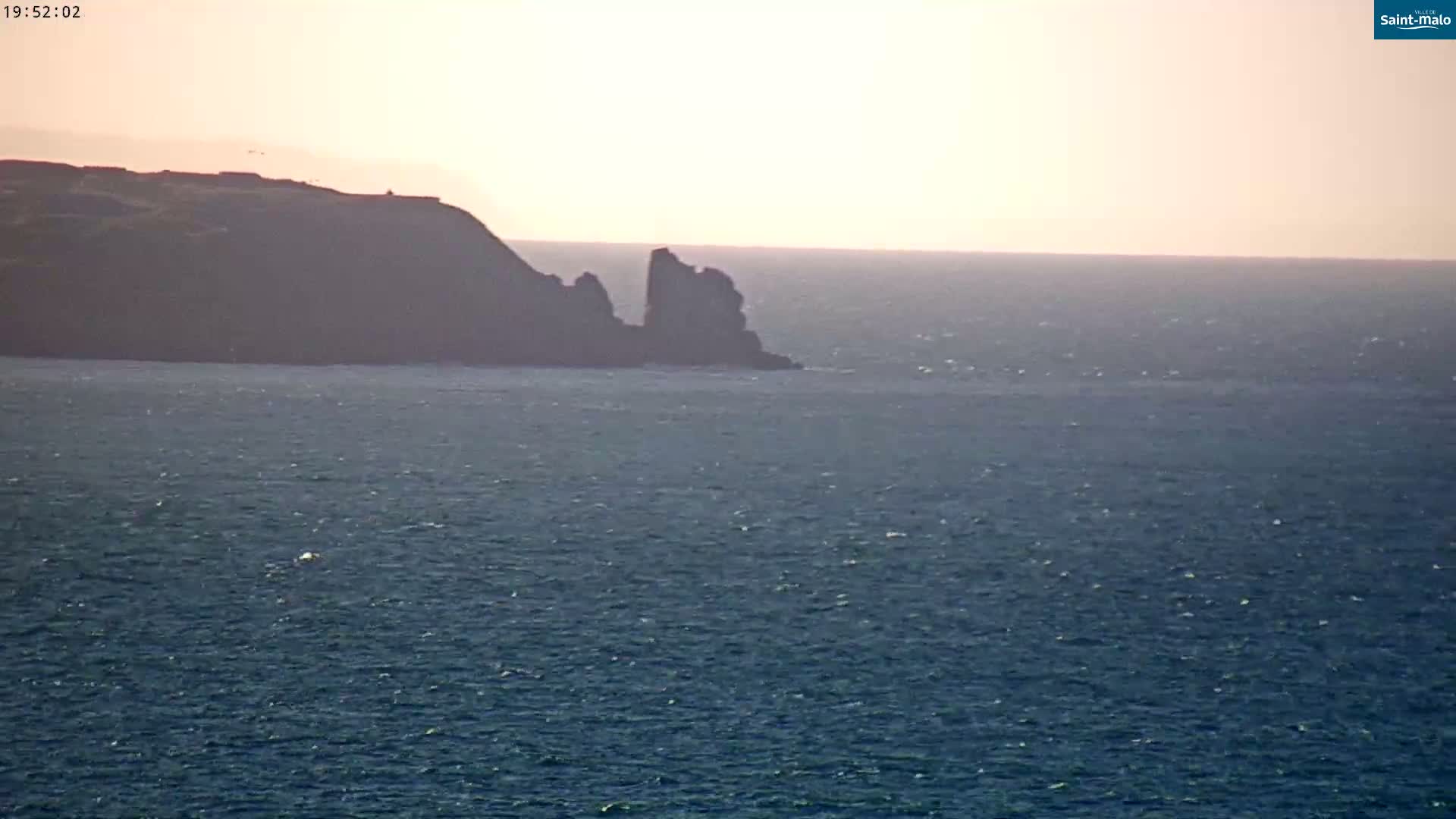 A dark, rocky headland juts into a calm, blue ocean under a hazy, light-pink sky.