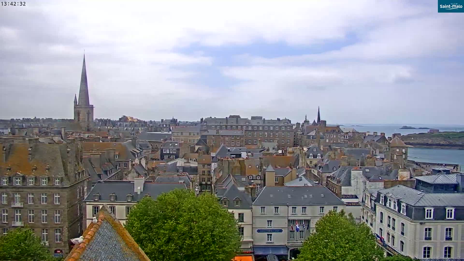 A sprawling cityscape of grey-roofed buildings under a mostly cloudy sky, with a tall church spire prominent in the center and a body of water visible in the distance.
