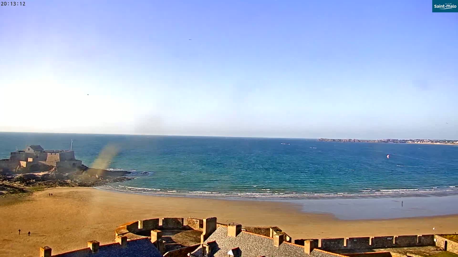 A sandy beach stretches out before a stone fortress overlooking a blue ocean under a clear sky, with a kitesurfer visible in the distance.