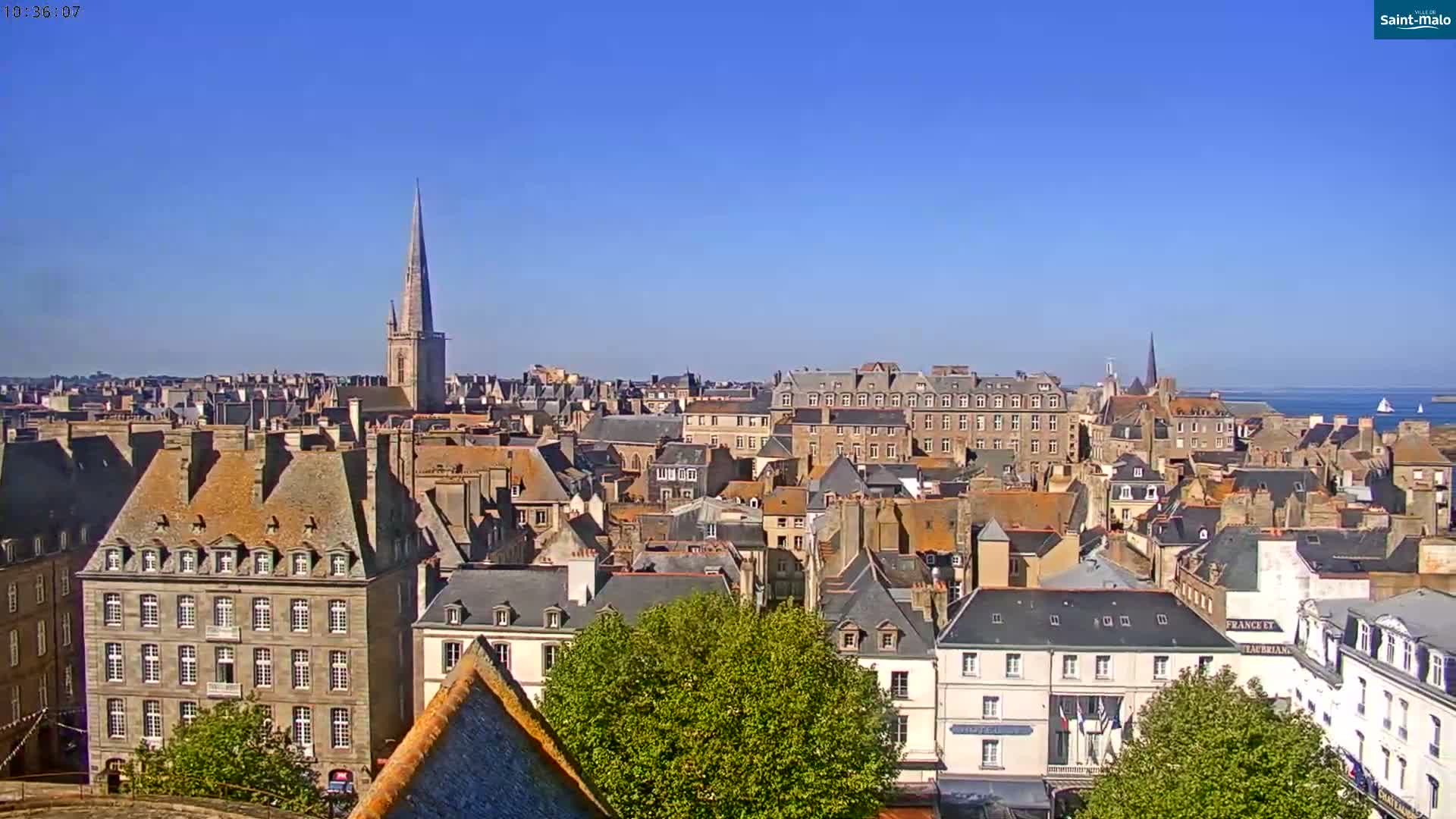 A panoramic view of a city with many tightly clustered buildings, a tall church spire, and a body of water in the distance under a clear blue sky.