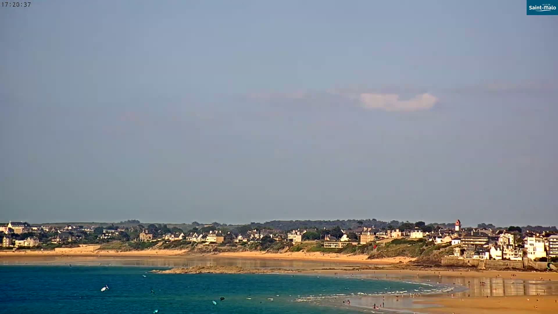 A mostly sunny day shows a coastal town with a sandy beach and calm, teal water.