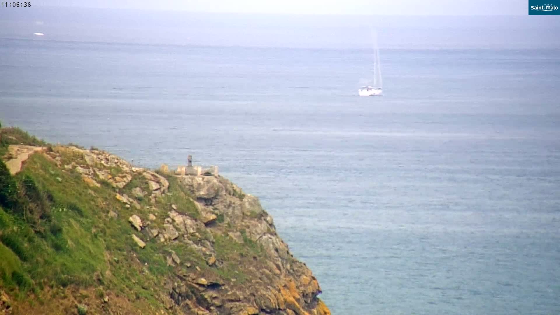 A sailboat is visible on a calm sea from a rocky, grassy clifftop where a small structure is situated.