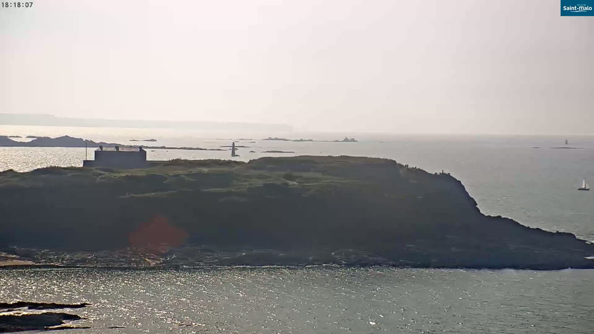 A hazy, sunny day reveals a rocky coastal area with a building on a small island, a body of water with a few sailboats, and distant landmasses.