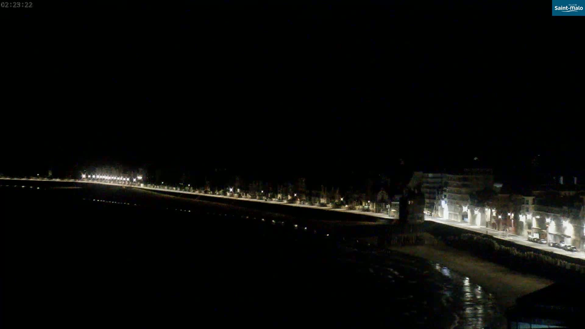 A nighttime, long-range view shows a dimly lit waterfront with buildings along the shoreline, partially obscured by darkness.
