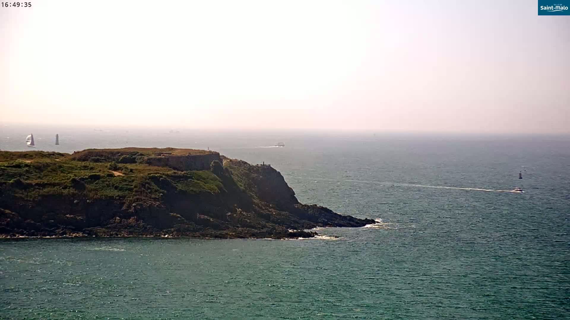 A rocky, green-covered headland juts into a hazy, blue ocean where several boats are visible in the distance under a bright, sunny sky.