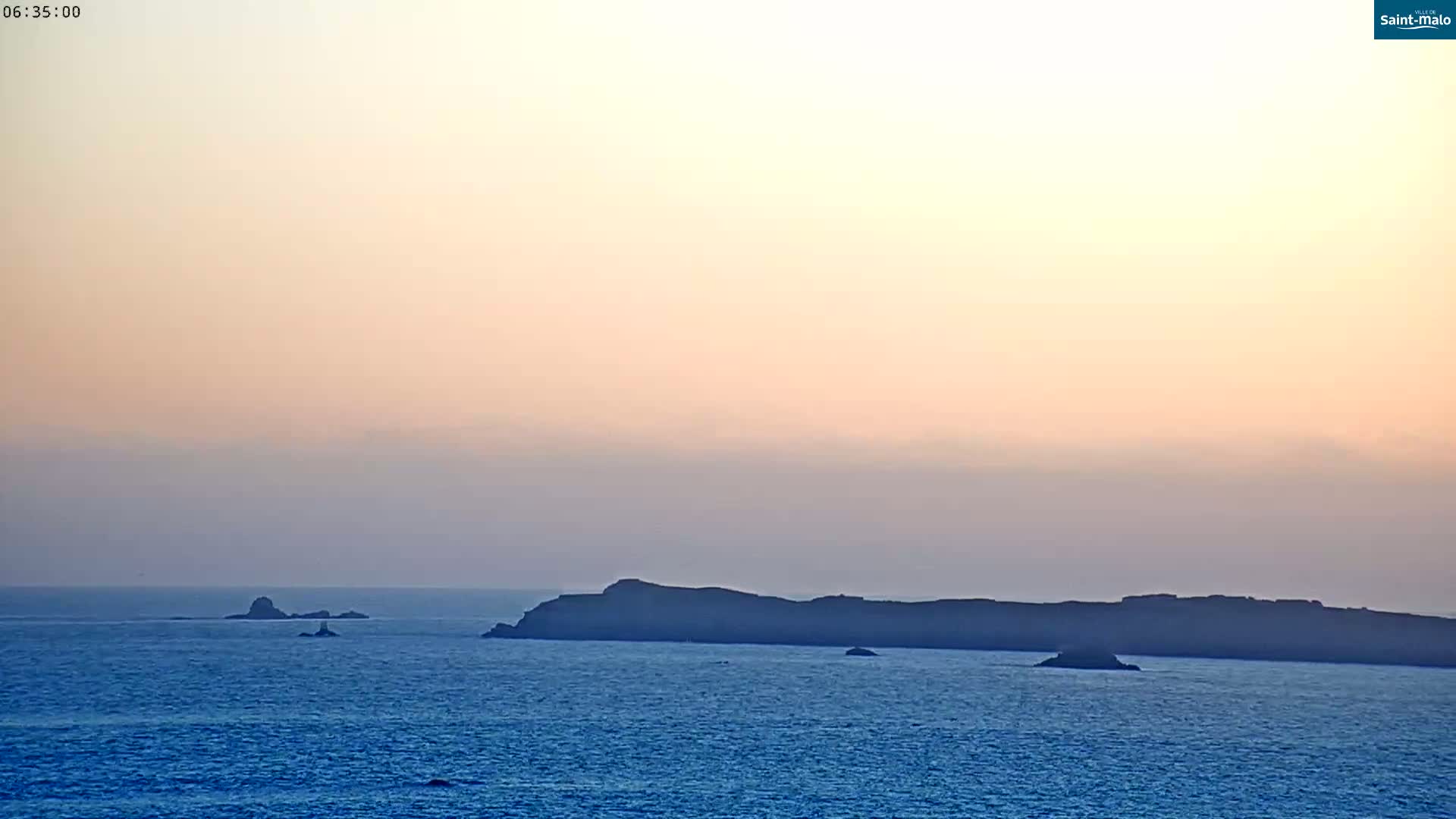 A calm ocean scene at sunset shows a few dark, small islands on the horizon under a pale orange and pink sky.