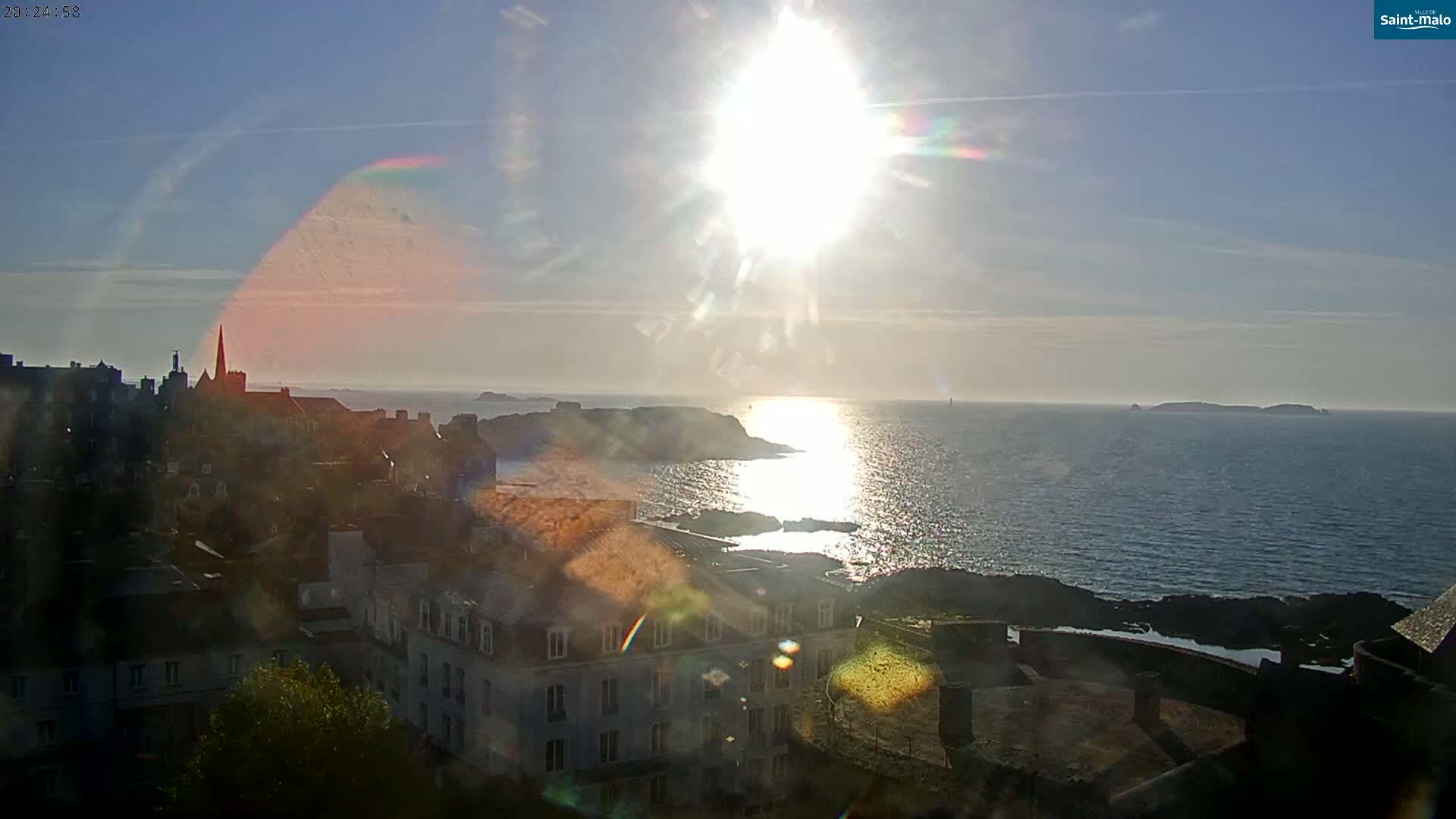 A bright, sunny day overlooks a coastal town with buildings and a church steeple,  a body of water reflecting the sun, and islands in the distance.