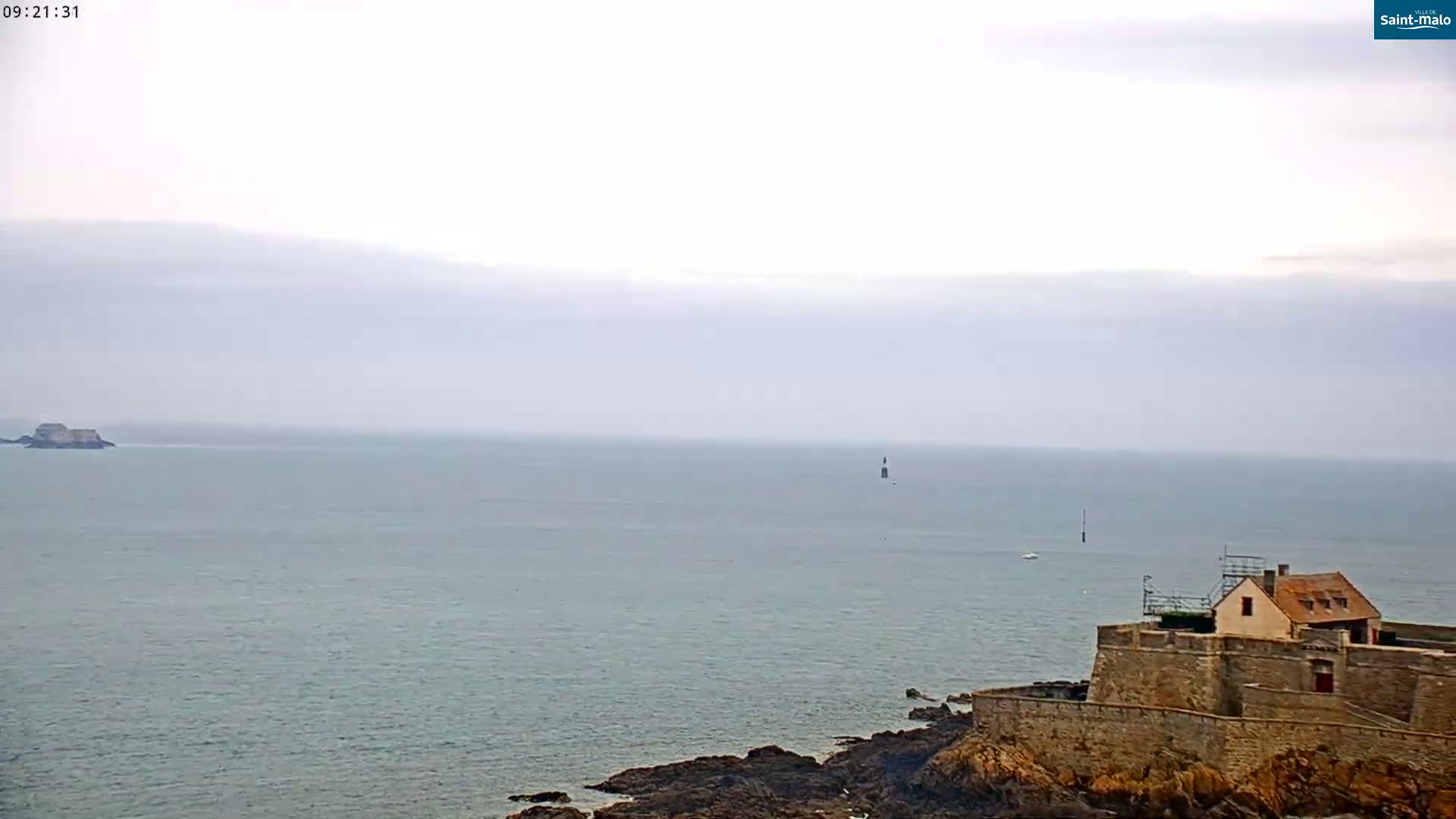 A stone building sits atop a rocky shoreline overlooking a calm, gray sea under a hazy sky.