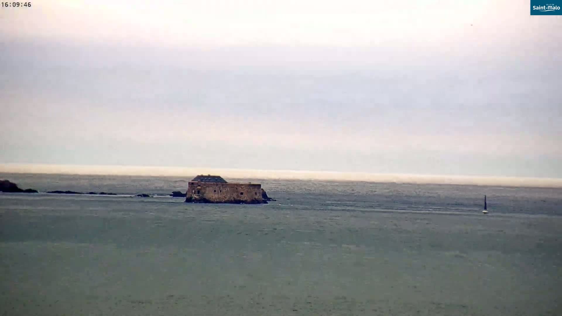 A stone fortress sits on a small island in a calm, hazy sea, with a sailboat visible in the distance.