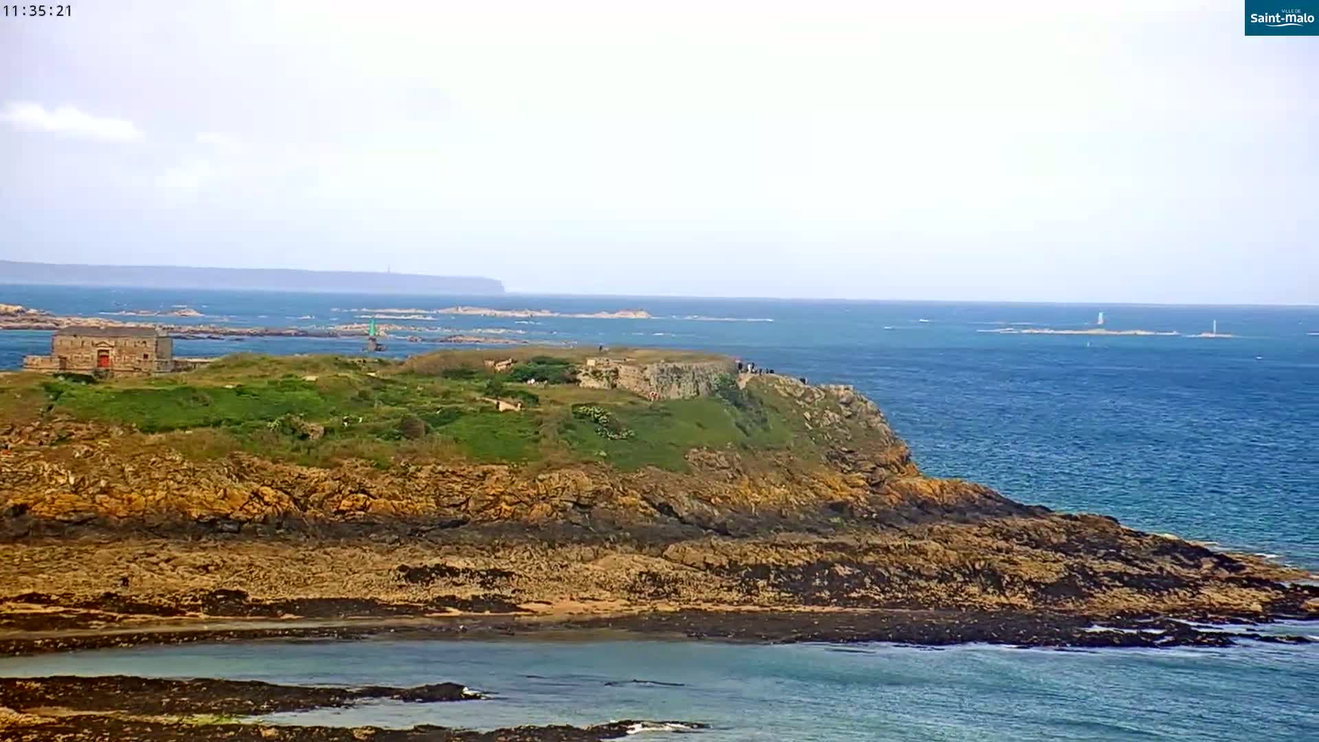 A rocky, grassy headland with a stone building overlooks a calm, blue ocean on a mostly cloudy day.
