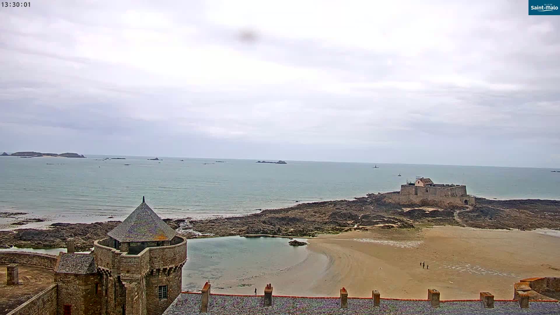 A coastal scene on an overcast day shows a stone tower overlooking a sandy beach, rocky coastline, a distant fortress, and several small islands in the ocean.